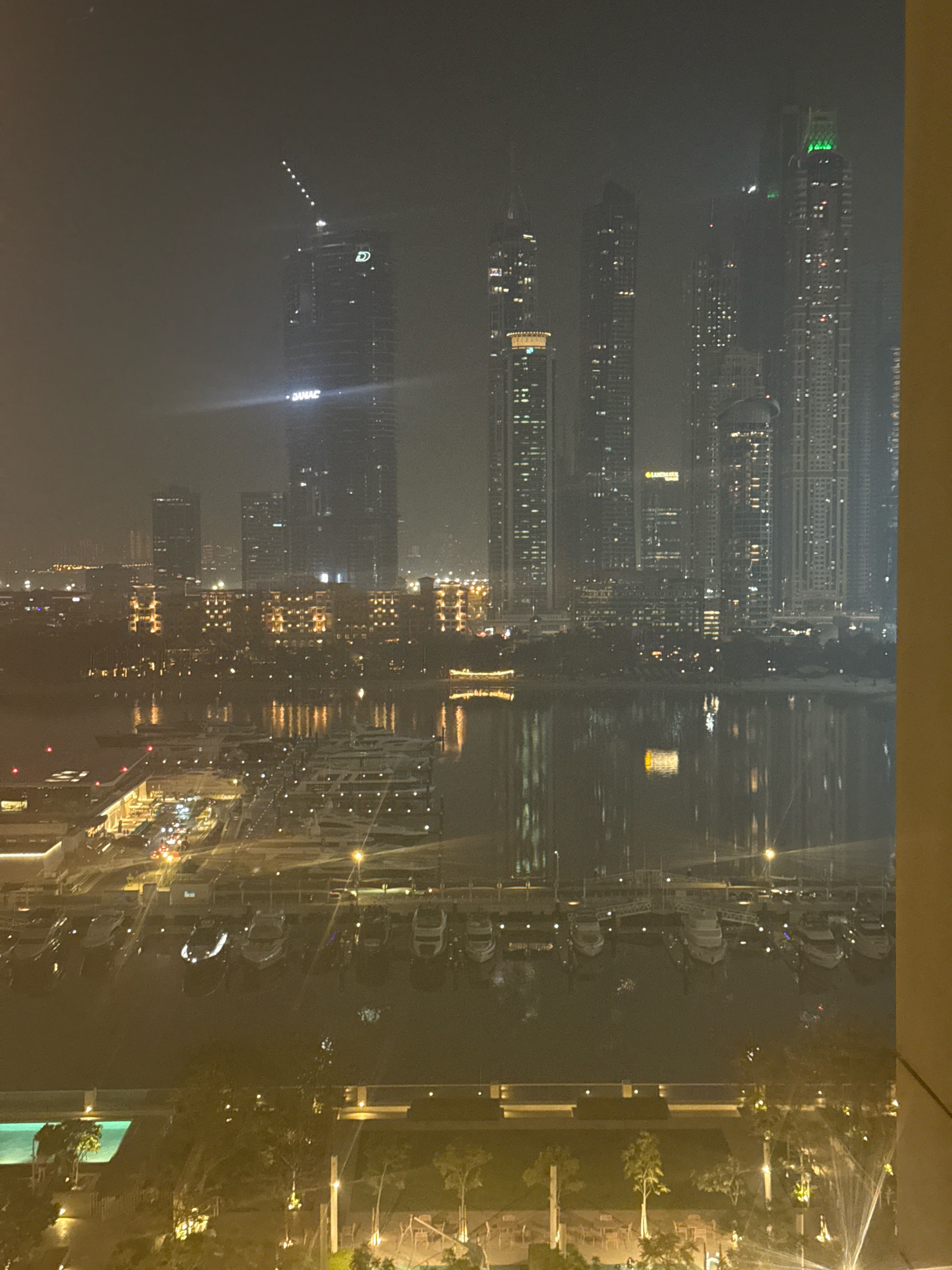 The image depicts a nighttime view of a waterfront in Dubai, United Arab Emirates, likely the Dubai Marina area, characterized by its dense concentration of modern skyscrapers and a marina filled with yachts. The sky is hazy, possibly due to fog or dust, obscuring the details of the buildings and creating a soft, diffused light.
In the foreground, a marina basin is visible, housing numerous docked yachts of various sizes. The lights from the yachts and the surrounding infrastructure are reflected in the calm water, creating shimmering patterns. Beyond the marina, a promenade or walkway is lined with trees and outdoor seating areas. A swimming pool with illuminated turquoise water is also visible on the left side of the image, suggesting a residential or hotel complex.
The midground is dominated by a cluster of towering skyscrapers, their windows illuminated, piercing the hazy night sky. These buildings showcase the characteristic modern architecture of Dubai, with sleek designs and varied heights. The lights of the city create a distinct urban glow against the dark backdrop. One prominent skyscraper in the center has a large illuminated logo that appears to read "DAMAC." Another tall building on the far right has a bright green illuminated top.
The background shows more of the urban landscape, with additional buildings and city lights extending into the distance. The overall impression is one of a vibrant, modern metropolis at night, with a strong emphasis on luxury waterfront living and impressive architectural achievements. The hazy atmosphere lends a slightly ethereal or moody quality to the scene. No people are clearly discernible in the image.