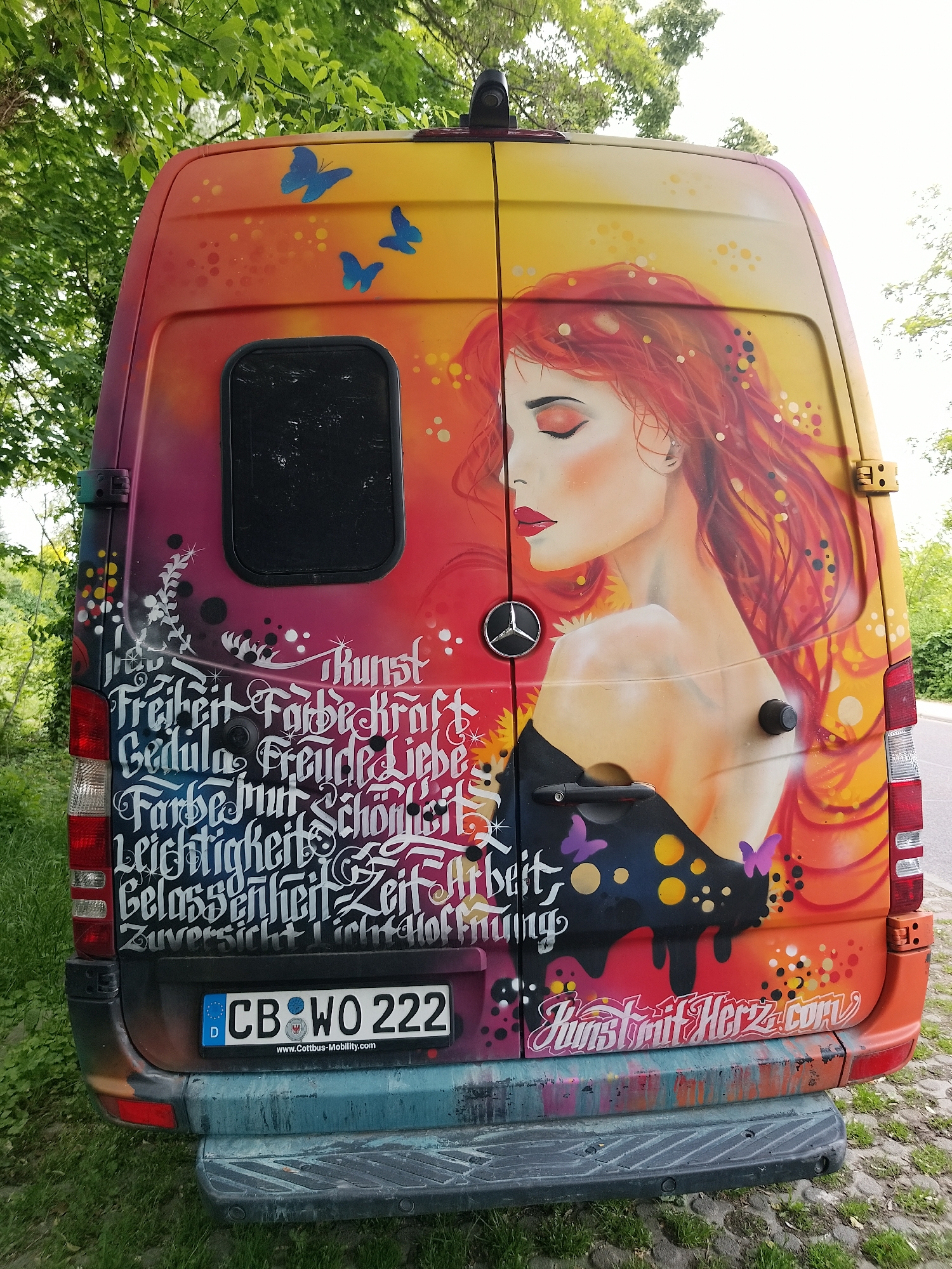 The image shows the rear of a brightly colored, artfully painted Mercedes-Benz van. The artwork includes a portrait of a woman with flowing red hair, butterflies, and stylized lettering. The van's license plate reads "CB WO 222." The text in the artwork includes the German words "Freiheit," "Farbe," "Liebe," and "Kunst." Below the artwork is the website address "KunstmitHerz.com." The van is parked outdoors, likely on the side of a road, with greenery visible in the background.