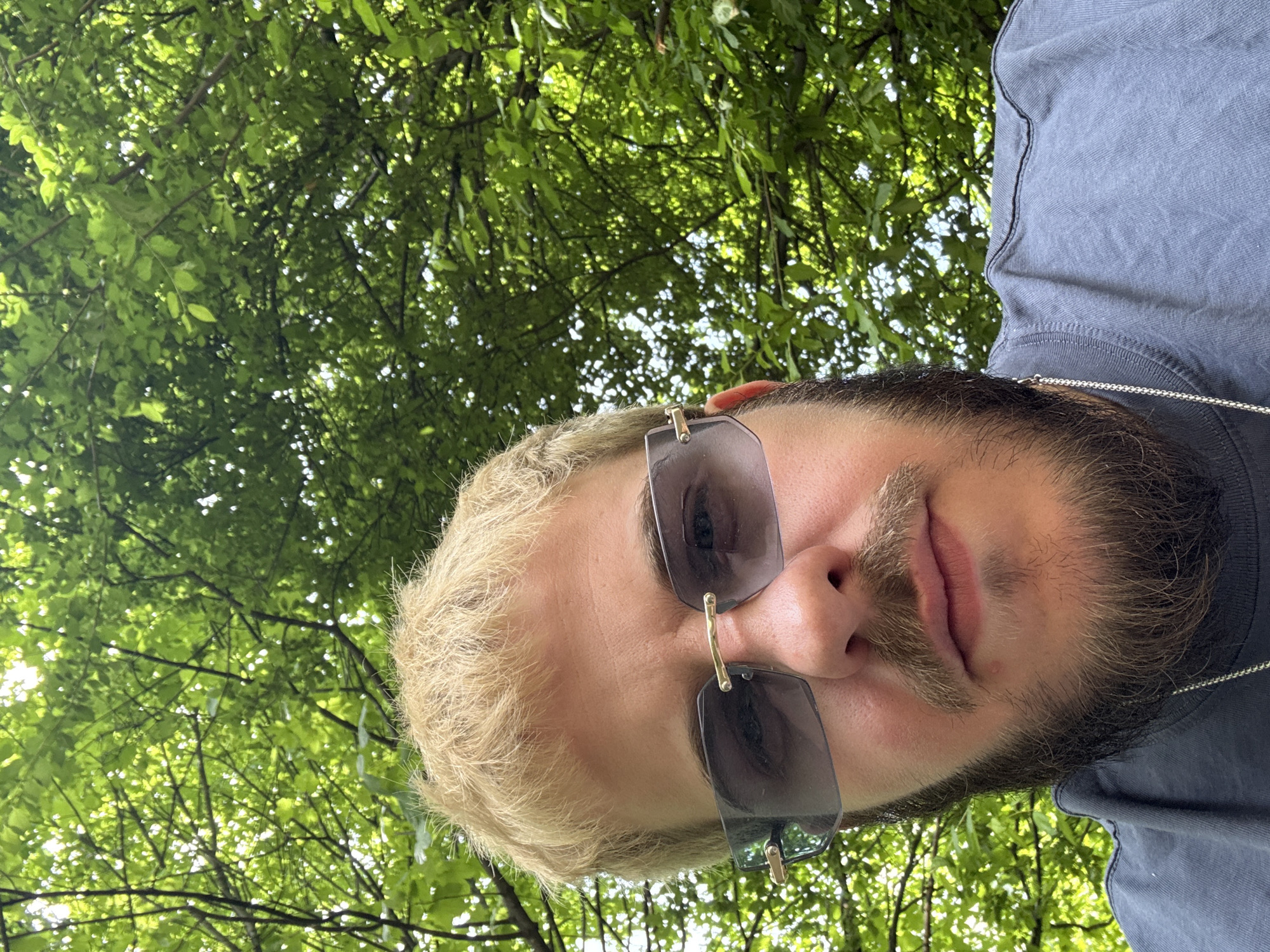 A man with blonde hair and a beard wearing sunglasses is looking up at the camera. Behind him, there are green trees. The man is wearing a grey t-shirt and a silver chain. The location is likely an outdoor park or wooded area, possibly in Sofia, Bulgaria.