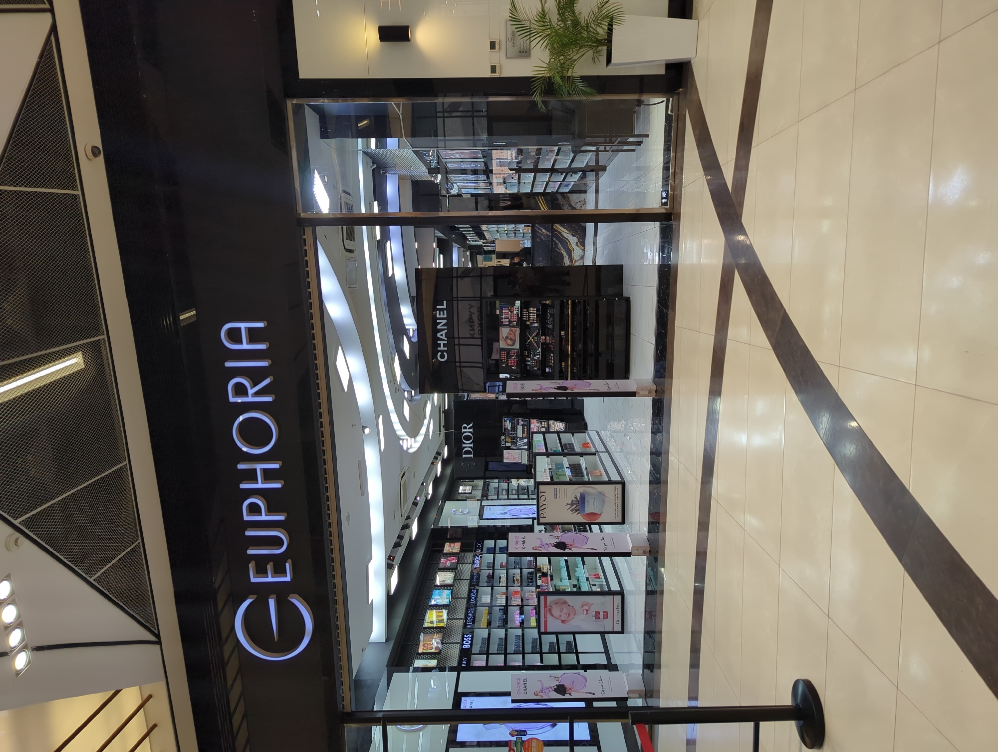 This image captures the front of a beauty store named "Euphoria" in Bishkek, Kyrgyzstan. The store features prominent displays of brands like Chanel, Dior, Boss, Versace, and Lancôme, with their logos visibly printed on signs and displays. The storefront is modern, with large glass windows allowing a clear view of the products inside. The store is located inside a mall, as indicated by the tiled floor and other storefronts visible in the reflection. The lighting is bright and even, highlighting the products and creating an inviting atmosphere. The overall ambiance suggests a well-organized, upscale retail environment. A small potted plant is placed near the entrance. There are also signs with product information on display in front of the store.