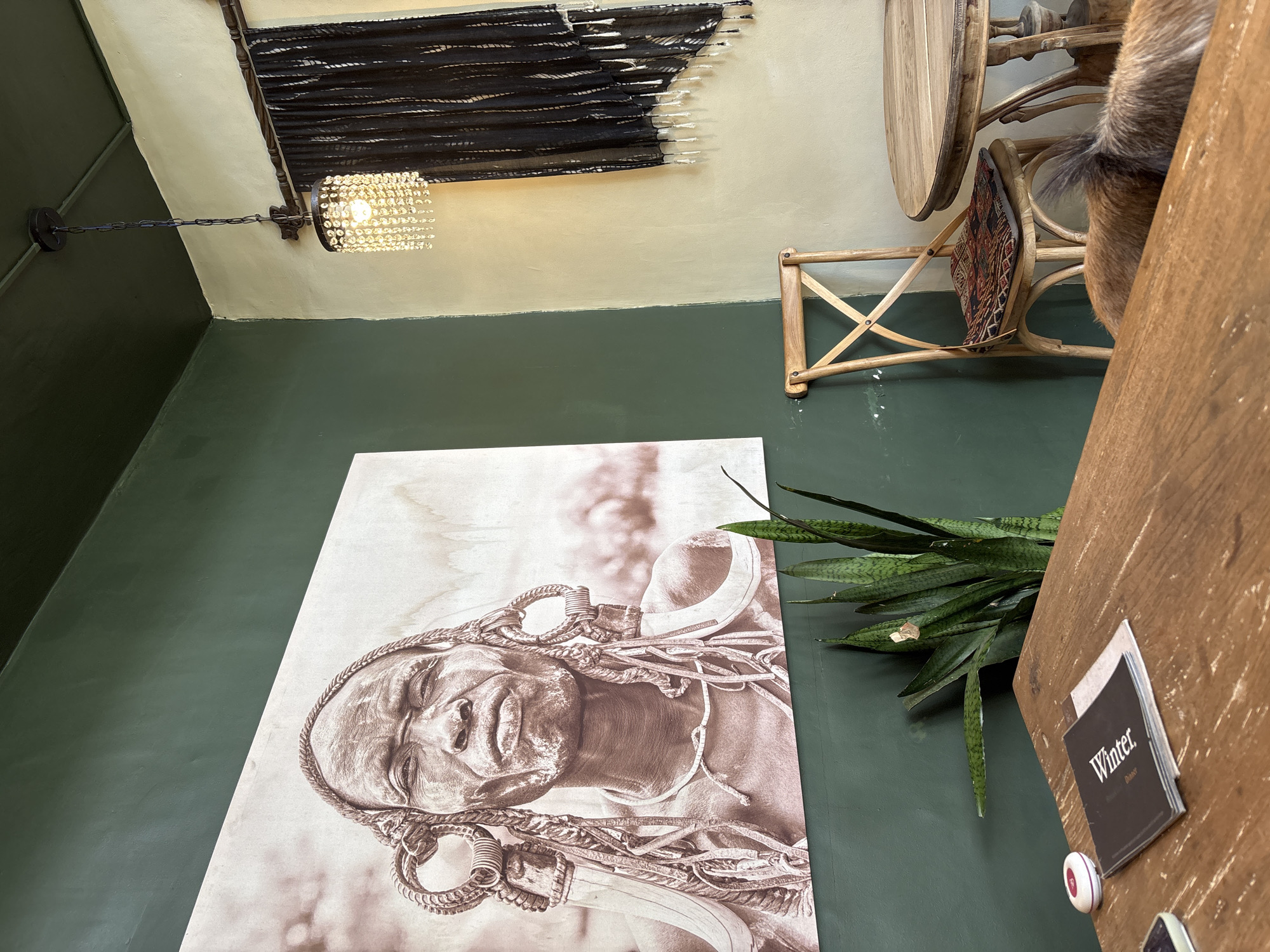 The image depicts an interior scene, possibly a cafe or restaurant, with distinct cultural elements. A large, sepia-toned portrait of an elder with elaborate earrings and headdress dominates the dark green wall on the left. To the right, a wall painted in a lighter, creamy yellow features a decorative black textile with fringe hanging near a chandelier with crystal accents. A wooden chair with an intricately patterned cushion sits between the two walls, positioned near a round wooden table. In the foreground, a section of a wooden table is visible, with a book titled "Winter" and a small white circular object on it. A snake plant is positioned between the large portrait and the table. Part of a small dog or other furry animal is visible in the lower right corner, resting near a table. The lighting suggests it is daytime, with natural light entering from an unseen source. The overall ambiance is one of artistic expression and cultural fusion. There is no visible indication of the city of Kuta Selatan, Indonesia.