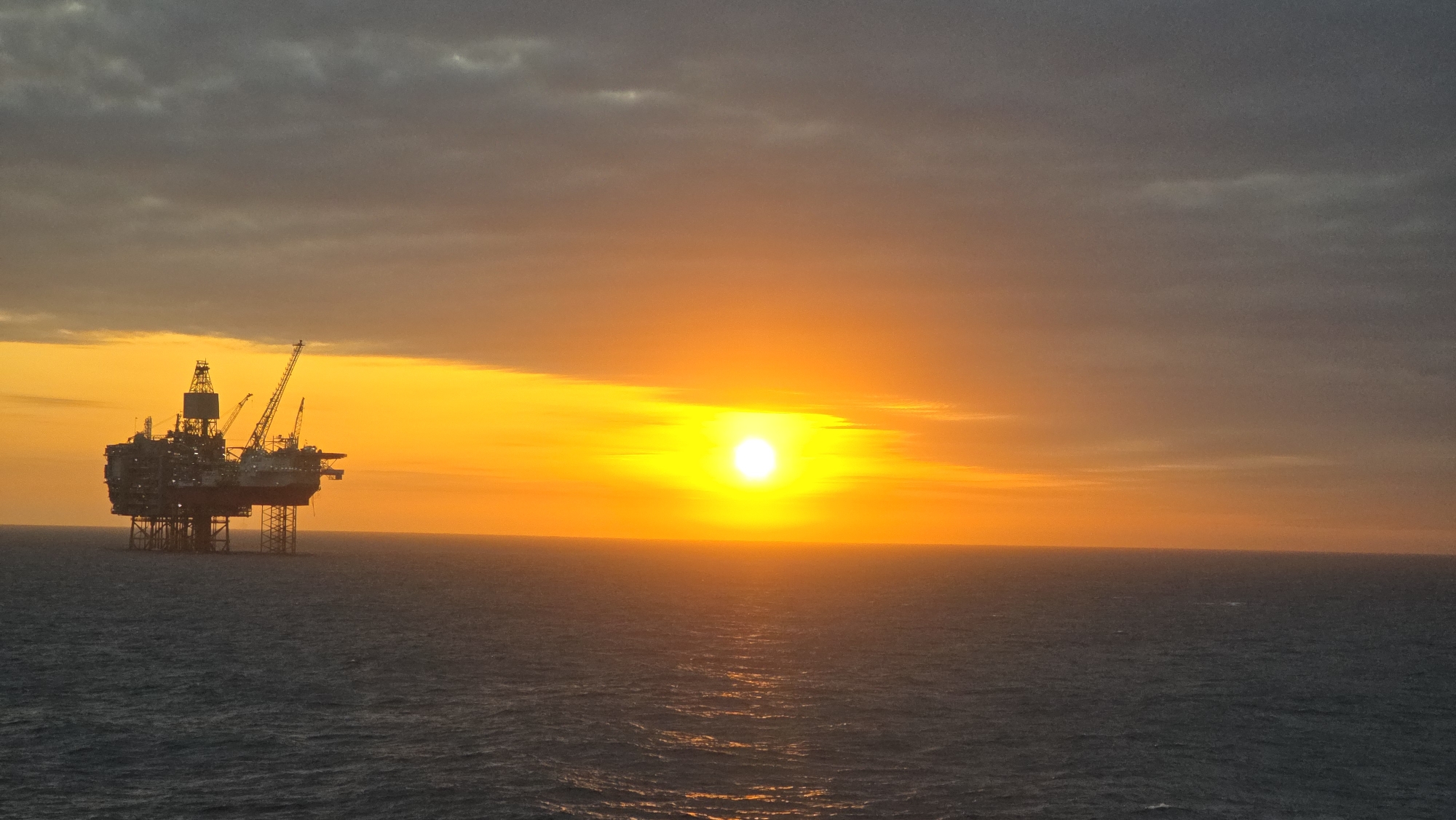 The image shows an offshore oil platform at sunset on the ocean. The platform, located on the left, is silhouetted against the bright orange and yellow sunset sky. A grey, cloudy sky overhangs the yellow sunset. The ocean is a dark blue-grey, with the sun reflecting on the water's surface. The location in the city of Randaberg, Norway is not evident from the image.