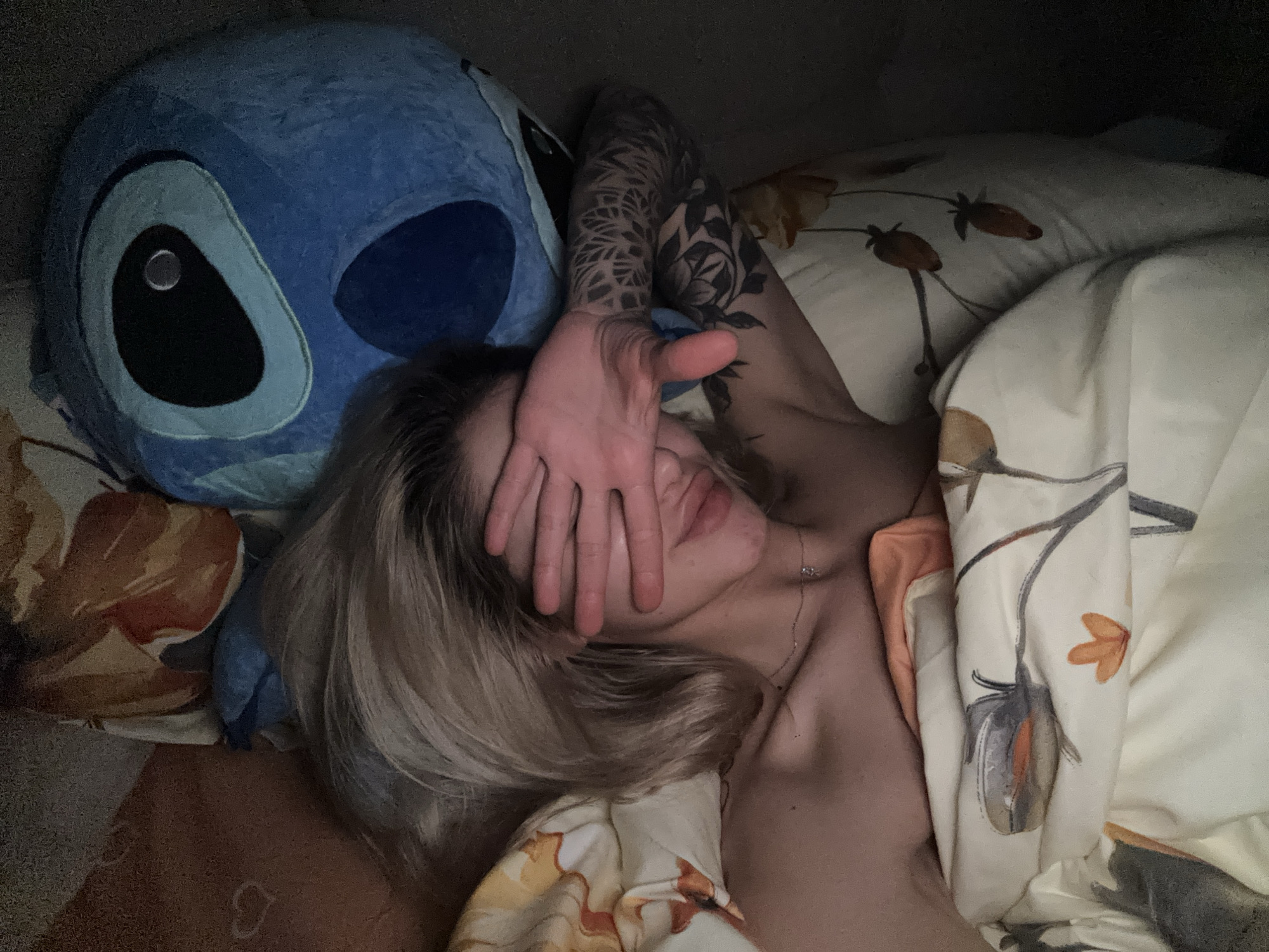 A person is lying in bed, covering their face with their hand. They have blonde hair and are wearing a necklace. Their left arm, which is visible, has a tattoo. Next to them is a large blue plush toy. The bedding has a floral pattern. The location is unknown.
