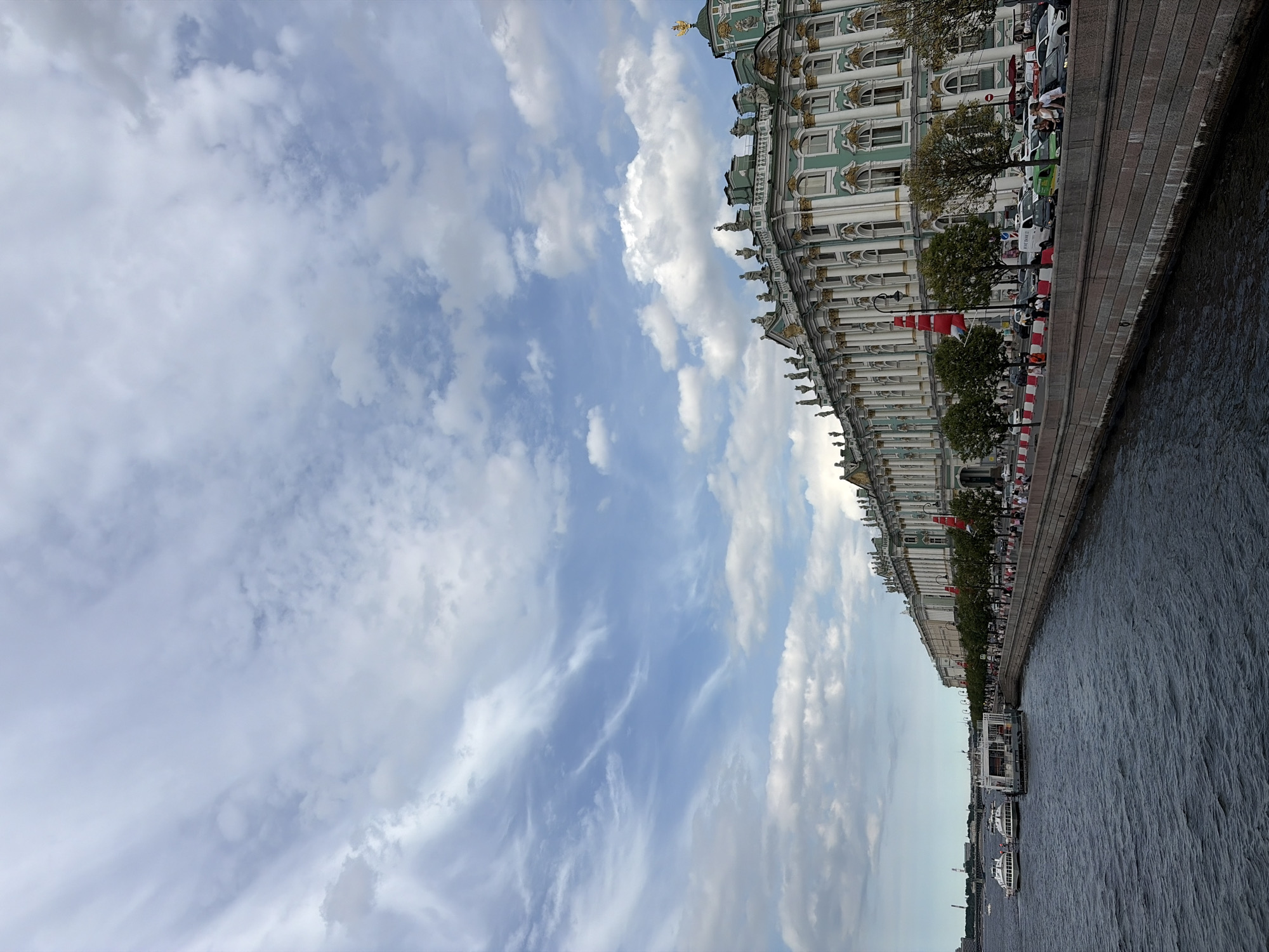 The image depicts a scenic view of a canal in Saint Petersburg, Russia, with the iconic Hermitage Museum visible along the waterfront. The perspective is from a slightly elevated viewpoint, looking across the water towards the grand building.
The Hermitage Museum is characterized by its distinctive light green facade, white columns, and ornate golden detailing. Its classical architecture stretches along the quay, punctuated by numerous windows and decorative elements. Along the riverbank in front of the museum, trees line the promenade, and several people are seen strolling or standing.
Several boats are docked or cruising on the dark, rippling water of the canal. These appear to be tour boats, suggesting a daytime scene where tourists are exploring the city's waterways. The water's surface is textured with small waves, reflecting the overcast sky.
The sky above is a mix of blue and white, with scattered clouds. Some clouds are wispy and streaky, indicating a dynamic sky, while others are more diffused. This suggests a partly cloudy day. The lighting is even, without harsh shadows, which points towards a time of day around midday or early afternoon, under cloudy or hazy conditions.
The overall atmosphere is one of historical grandeur and leisurely tourism. The scene is tranquil, with the gentle movement of the water and the distant presence of people. The orientation of the image is vertical, capturing a wide expanse of the sky and the long facade of the museum. No visible text is present in the image.