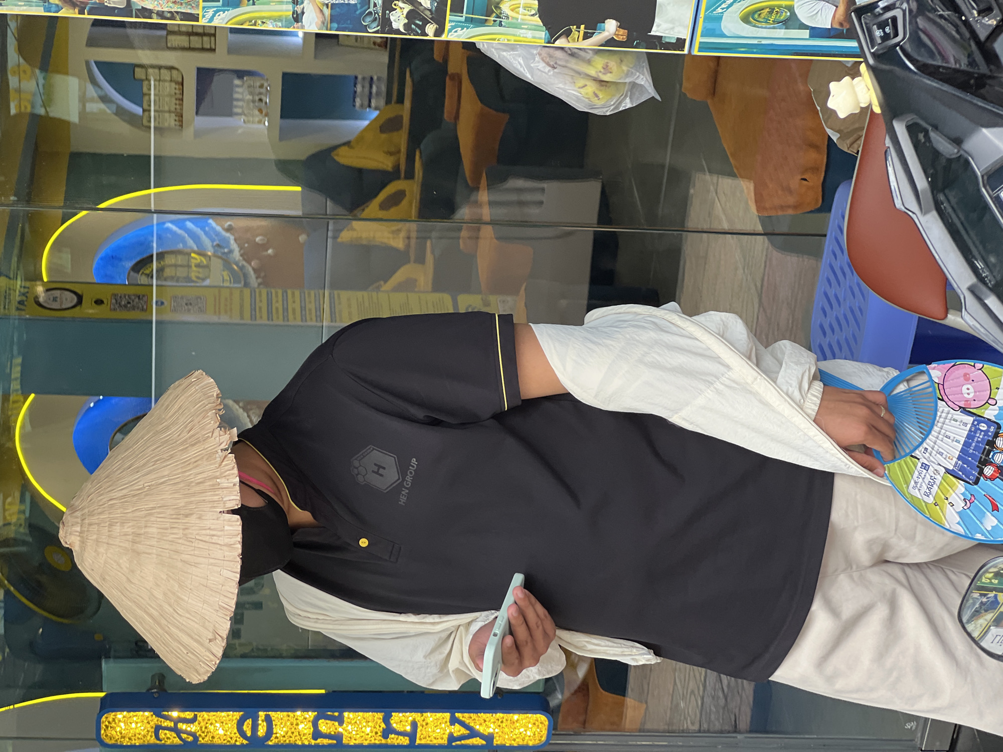 The image depicts a person in Nha Trang, Vietnam, standing in front of a shop window. The person is wearing a conical hat, a black polo shirt with the "HEN GROUP" logo, a white jacket draped over their shoulders, and light-colored shorts. They are holding a smartphone in their right hand. The shop window reflects the interior, which appears to be a spa or massage parlor with reclining chairs and ambient lighting. The background also shows various signs and posters, typical of a commercial street in Vietnam. The overall scene is a candid street-style shot, capturing a moment in a bustling urban environment.