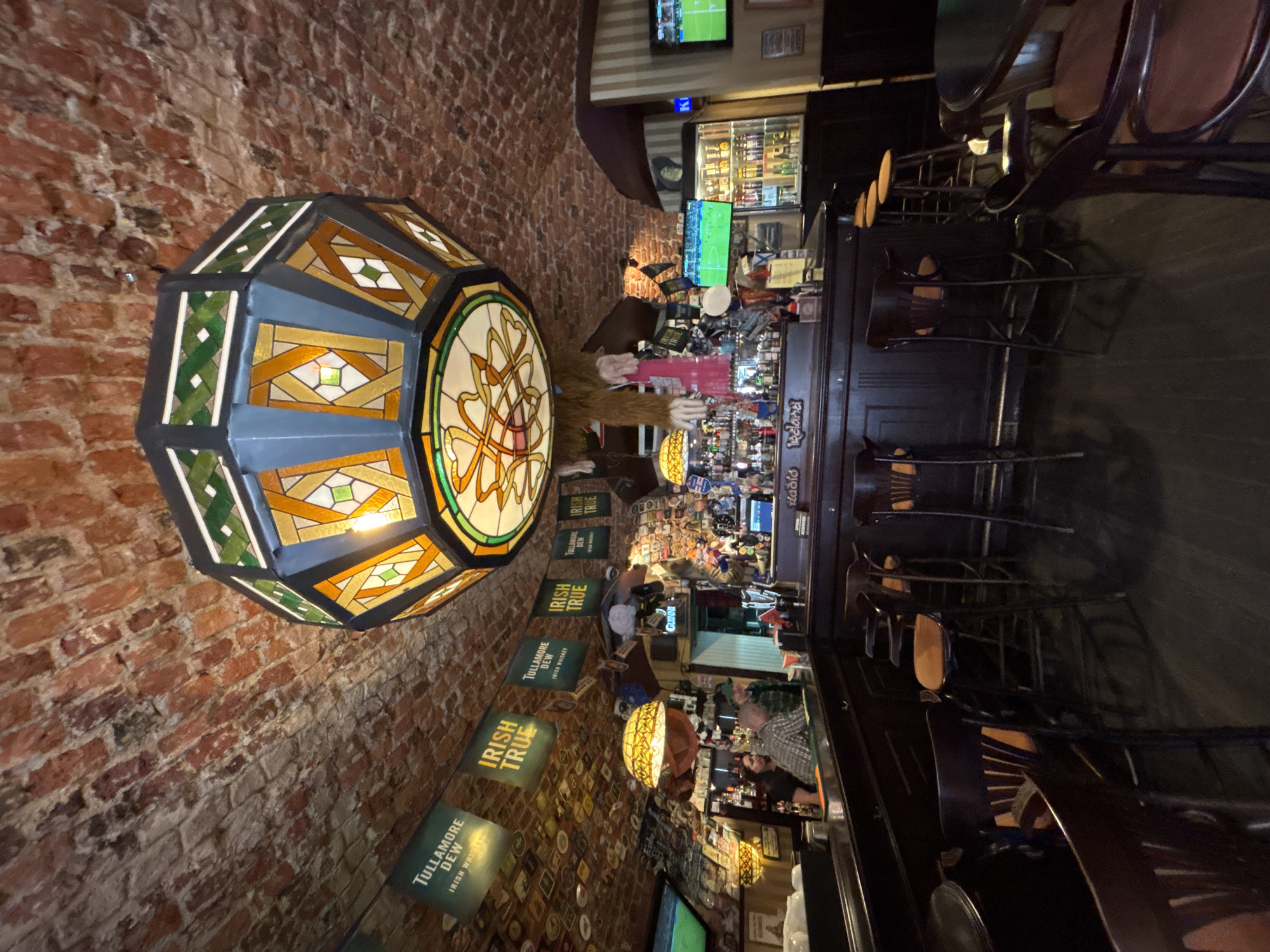 This image depicts the interior of a dimly lit Irish pub in Saint Petersburg, Russia. The scene is characterized by exposed brick walls and dark wooden furnishings, creating a cozy and traditional atmosphere. A prominent stained-glass hanging lamp with geometric patterns in shades of amber, green, and blue hangs from the ceiling on the left side of the frame, illuminating the area.
Several promotional banners for "Tullamore Dew Irish Whiskey" and "Irish True" are visible on the brick wall, suggesting a focus on Irish beverages and culture. In the background, a long bar area is stocked with various bottles of alcohol, and several bar stools with brown leather seats are arranged along it. A few patrons are present, including a person sitting at the bar, facing away from the camera, and another individual partially visible behind the bar.
Various decorative items, such as signs, bottles, and what appears to be a Guinness advertisement, are displayed on shelves behind the bar. Two television screens are also visible, one showing a sports game. The overall impression is of a lively pub setting, likely in the evening, with patrons enjoying drinks and socializing. The presence of Russian text on one of the signs ("Hadid ireland") reinforces the location context.