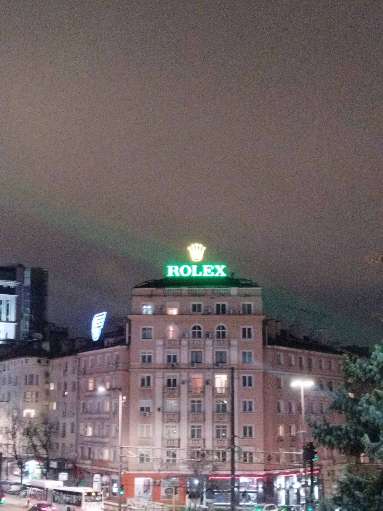 The image depicts a nighttime urban scene in Sofia, Bulgaria, centered on a multi-story building located at the intersection of Tsar Osvoboditel Blvd and Georgi Rakovski Street, near Narodno Sabranie Square. This light-colored structure is topped with a prominent, illuminated green "ROLEX" sign and crown logo. To the left, another multi-story building features a blue, illuminated wing-shaped sign. The street level below shows active vehicular traffic, including a white public transport bus and several cars with illuminated lights, operating under green traffic signals. Numerous windows within the buildings are illuminated, alongside bright streetlights and shopfronts at ground level. The sky is dark and overcast, exhibiting a slight greenish cast from light pollution. A section of an evergreen tree is visible on the right side of the frame.