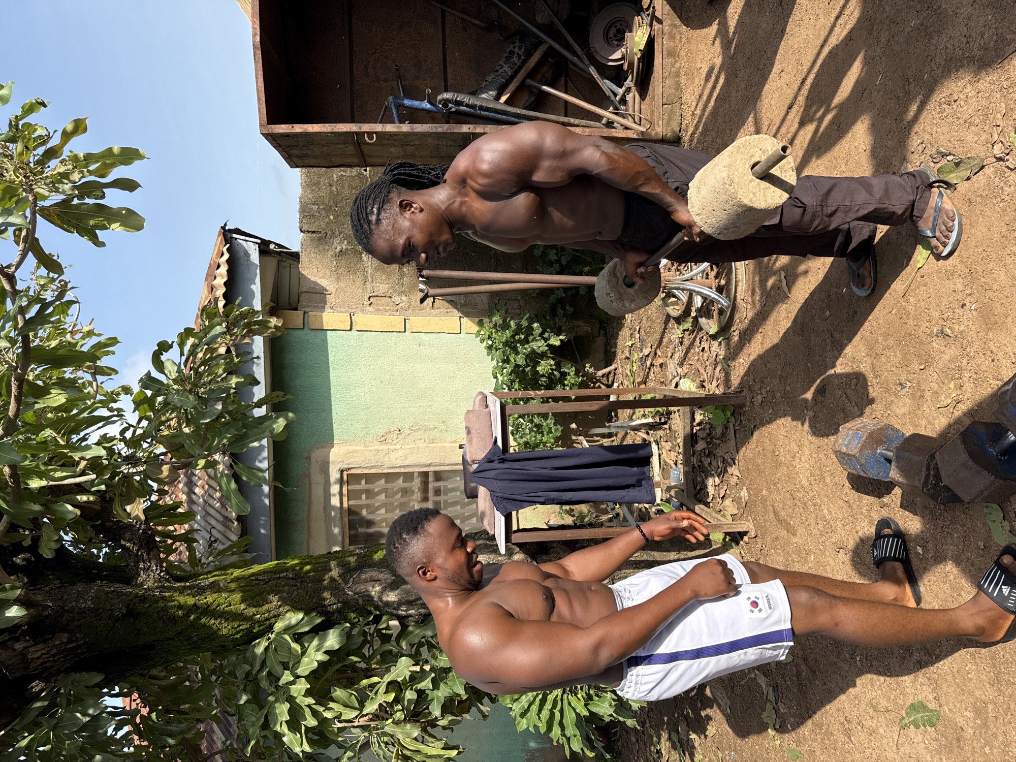 Two shirtless men are exercising outdoors in Akwanga, Nigeria. The man on the left, wearing white shorts with blue stripes, stands beside a metal rack with a dark blue garment draped over it. He is looking down and appears to be wiping sweat from his face. The man on the right, wearing dark pants, is holding a dumbbell made of concrete. He is looking towards the man on the left. Both men have muscular physiques. The environment is dusty and appears to be a yard with a house in the background. A large tree with green leaves is to the left of the frame. The sky is clear and blue.