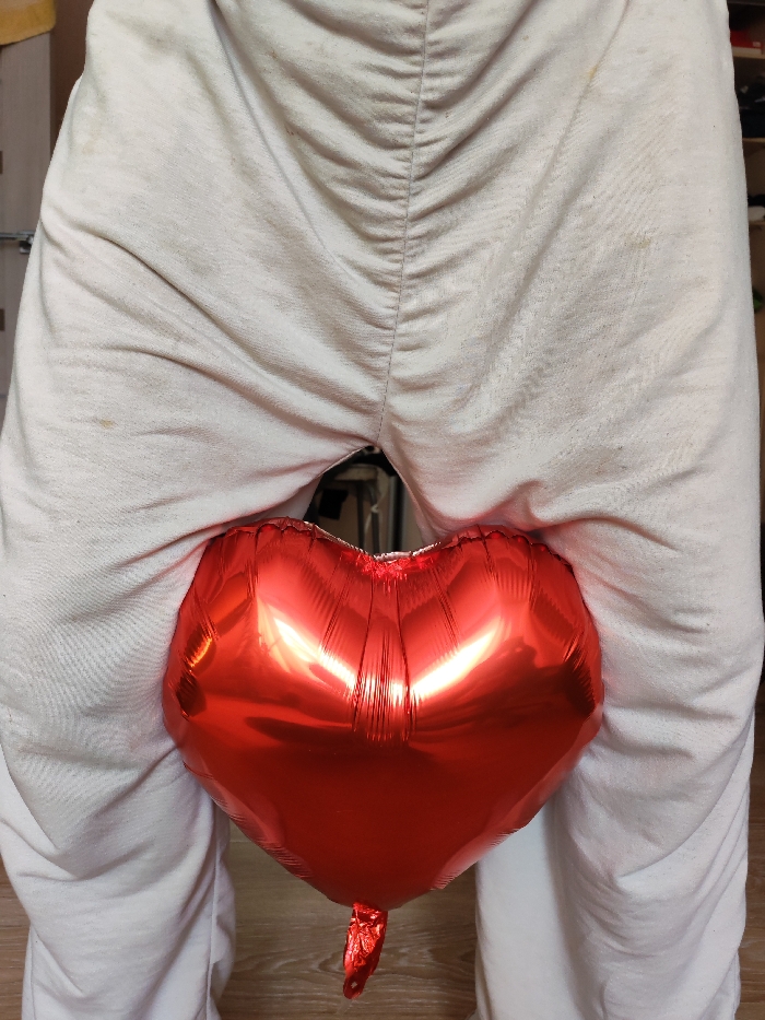 A partial view of a person standing indoors in Chaikovsky, Russia, is depicted. The lower body of the individual is visible, wearing light-colored, possibly off-white, creased fabric trousers or sweatpants that exhibit minor surface discoloration. A vertical seam or gathered fabric is prominent along the rear midline of the garment. A single red, inflated, heart-shaped foil balloon is positioned between the person's upper thighs, obscuring the crotch area. The balloon's reflective surface shows highlights from an unseen light source. The person's feet are not visible, but a light-colored wooden floor is present at the bottom of the frame. The background beyond the person's legs is out of focus, showing indistinct indoor elements.