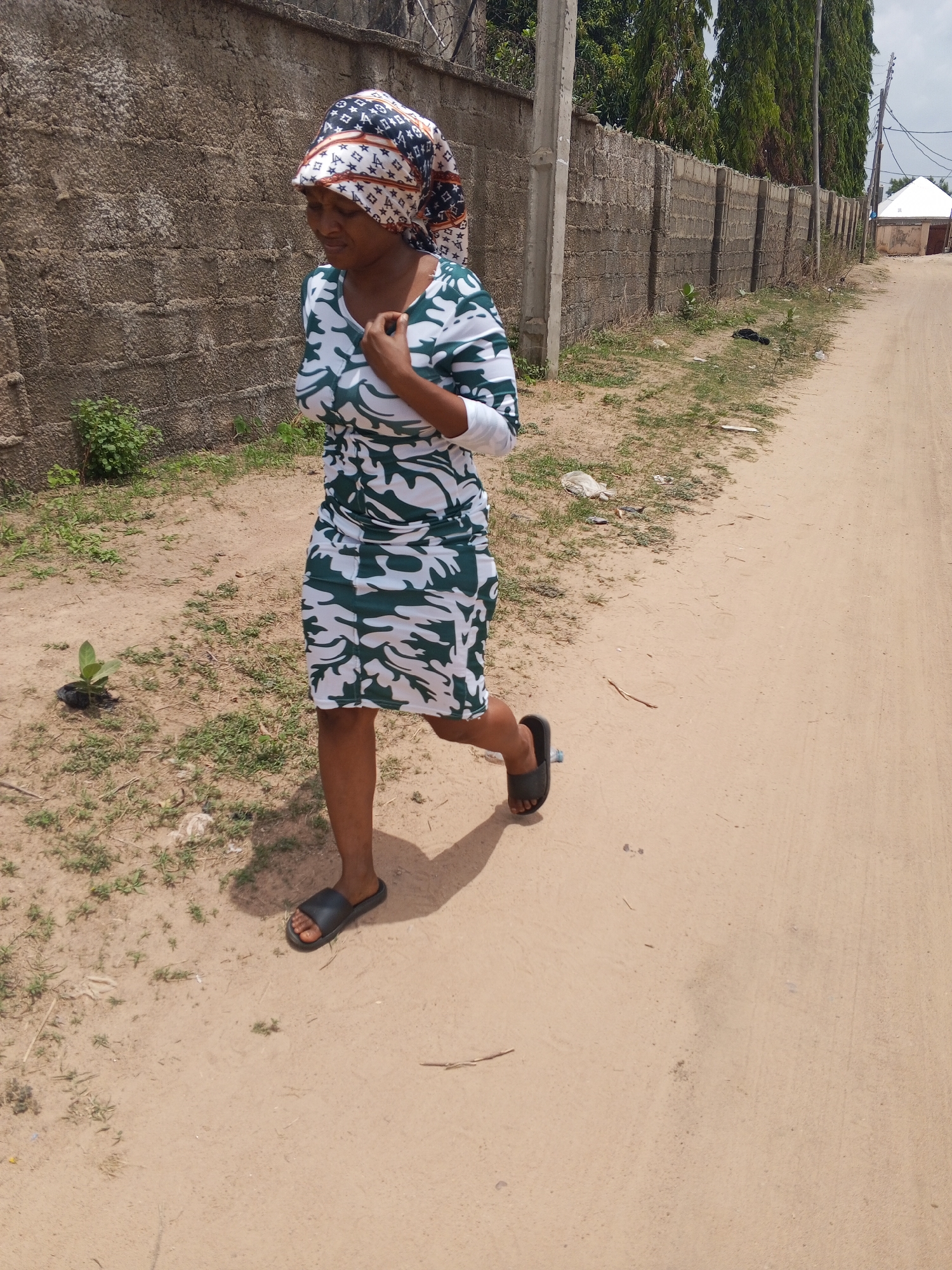 A woman is walking along a dirt road in Otokiti, Nigeria. She is wearing a patterned headscarf, a white and green dress with long sleeves, and black sandals. She is carrying her right hand to her chest. To her left is a gray brick wall. To her right is the dirt road, with some vegetation between the road and another wall in the distance. In the distance, a white building is visible. The sky is overcast.