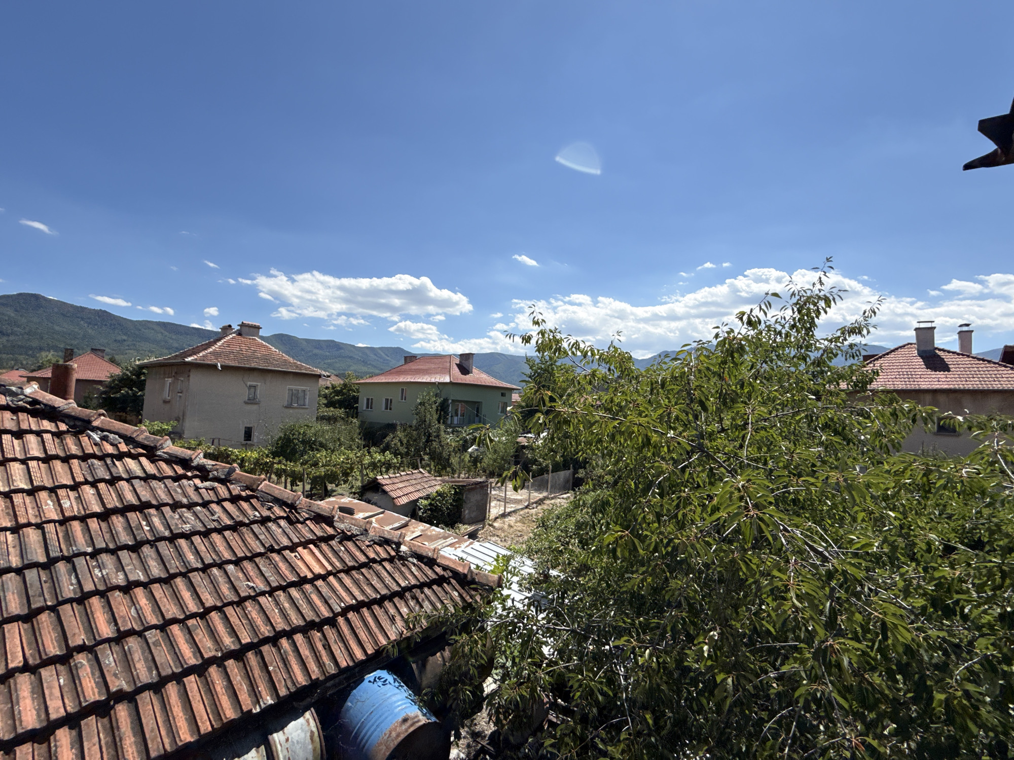 The image is an outdoor, eye-level shot looking out over a residential area in the city of Varshets, Bulgaria. The foreground is dominated by a close-up of a terracotta tiled roof, showing wear and tear. To the left and slightly below the roofline, two blue barrels are partially visible. In the mid-ground, several houses with tiled roofs are scattered among lush green trees and vegetation. One house in the mid-ground has a red tiled roof and a chimney. Further back, another larger house with a red tiled roof and two chimneys is visible. The background consists of rolling, tree-covered hills and mountains under a bright blue sky with scattered white clouds. The lighting suggests it is daytime, likely mid-morning or afternoon, with clear visibility. There are no people or animals in the image. The scene appears to be a quiet, rural or semi-rural setting within the city.