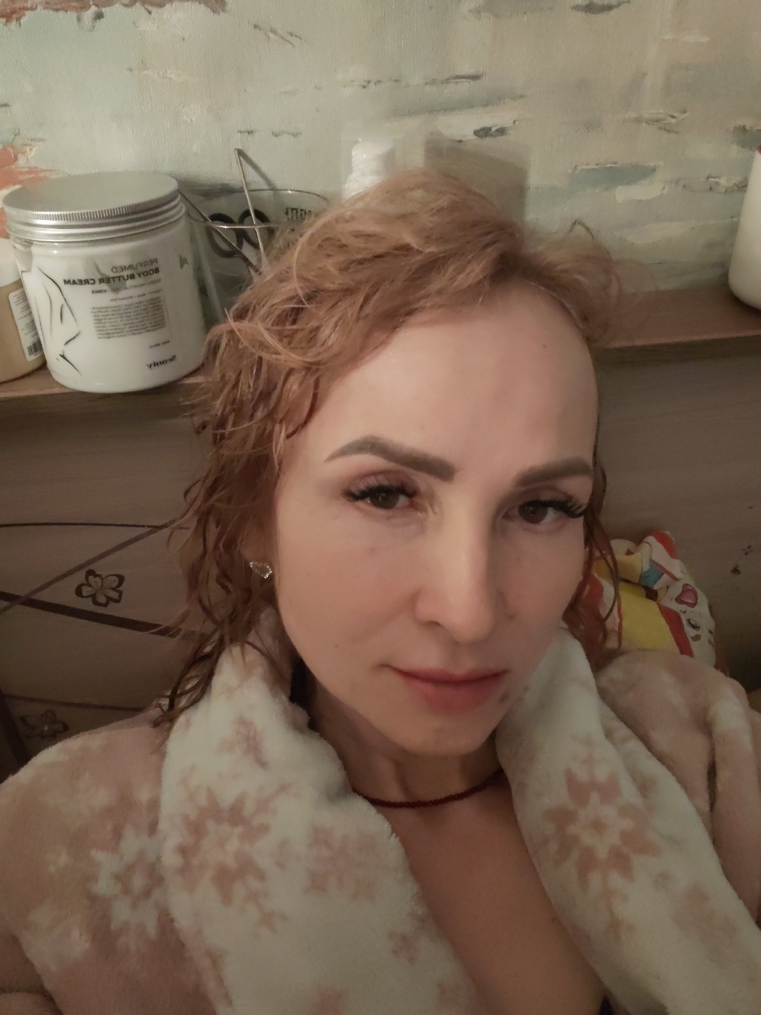 A woman with light brown, wet-looking curly hair takes a close-up selfie indoors. She has defined eyebrows, long dark eyelashes, and a neutral expression, looking directly at the camera. A thin, dark red beaded necklace is visible around her neck. She is wearing a fluffy, light pink and white patterned robe or blanket with snowflake designs.
The background shows a cluttered shelf or surface. To her left, a large white jar with a silver lid is visible. Inverted text on the jar reads "PERFUMED BODY BUTTER CREAM" and "BEAUTY". Next to it, a clear glass contains a metallic object. The wall behind her features a textured or painted pattern in light blue and white, resembling abstract brushstrokes. A colorful, possibly plush, object is partially visible to her right. The scene is lit by artificial light, suggesting an indoor setting, potentially in the evening. The context for the location is Chaikovsky, Russia.