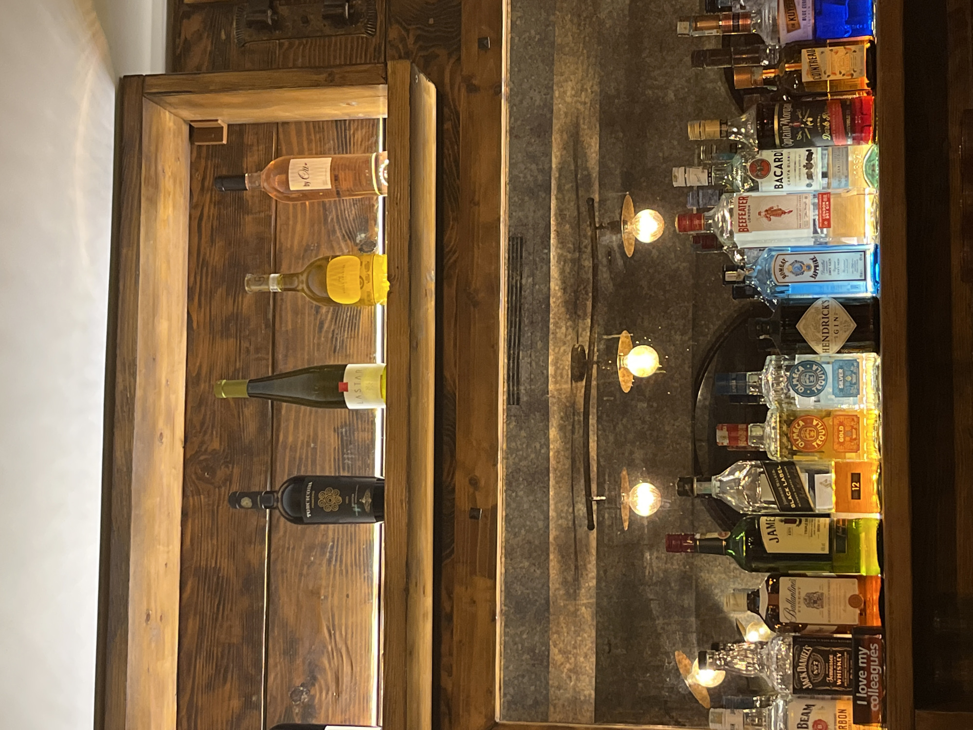 The image shows a liquor cabinet with various bottles of wine and spirits. On the left side of the cabinet, bottles of wine are horizontally stacked in a wooden rack. The bottles include a rose, a white wine, a bottle of green wine, and a bottle of red wine. The right side of the cabinet displays a collection of alcohol bottles arranged in a tiered fashion. The bottles include Hendrick's Gin, Bombay Sapphire, Beefeater Gin, Bacardi, Captain Morgan, Cointreau, Kuype Blue Curacao, Johnnie Walker Black Label, and Jack Daniel's Whiskey. Small, warm-toned light fixtures are spaced around the perimeter of the bottle display on the right side. The location in Budva, Montenegro, is unknown.