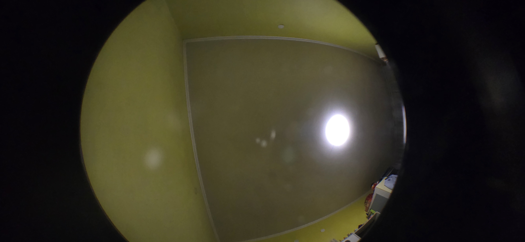 The image appears to be taken through a fisheye lens, possibly a peephole or similar device, showing an interior room. The walls and ceiling are painted in shades of yellow. The room is illuminated by a bright light source, presumably a ceiling fixture. The composition suggests that the image was taken from a low vantage point, possibly from a door or a small opening. The edge of the room shows possible furniture or items. It is not possible to determine the location in Sofia, Bulgaria, based on this image.