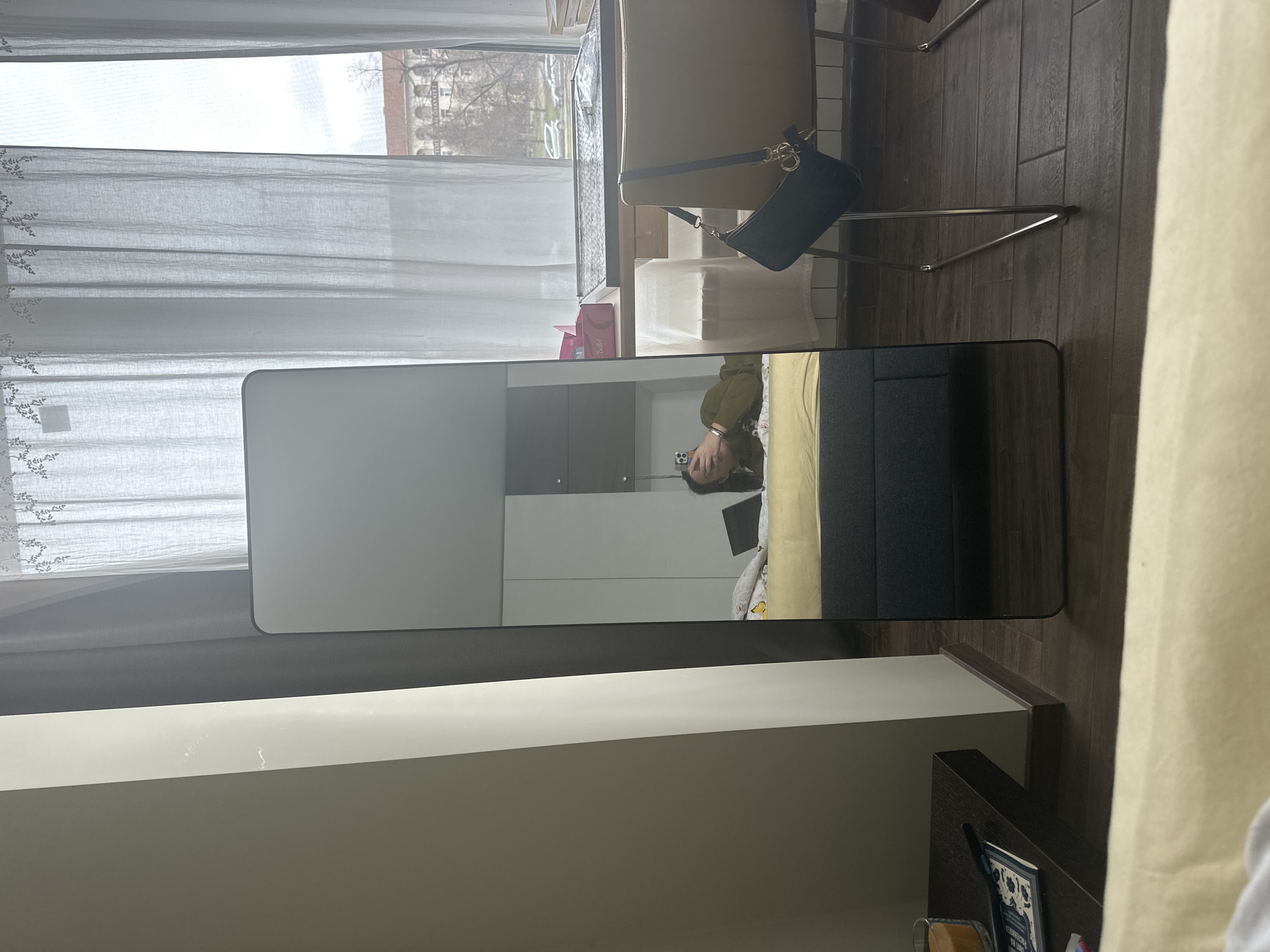 This is an indoor shot in what appears to be a bedroom. A full-length mirror reflects a person lying on a bed and holding a mobile phone. The bed has yellow bedding and a gray headboard. The room contains a desk with a white surface and a dark brown accent piece. A handbag hangs on the back of a beige chair positioned near a window. The window is covered with sheer white curtains and overlooks a cityscape, which appears to be in Sofia, Bulgaria, evidenced by the architectural style of the buildings and the leafless trees characteristic of the local climate.