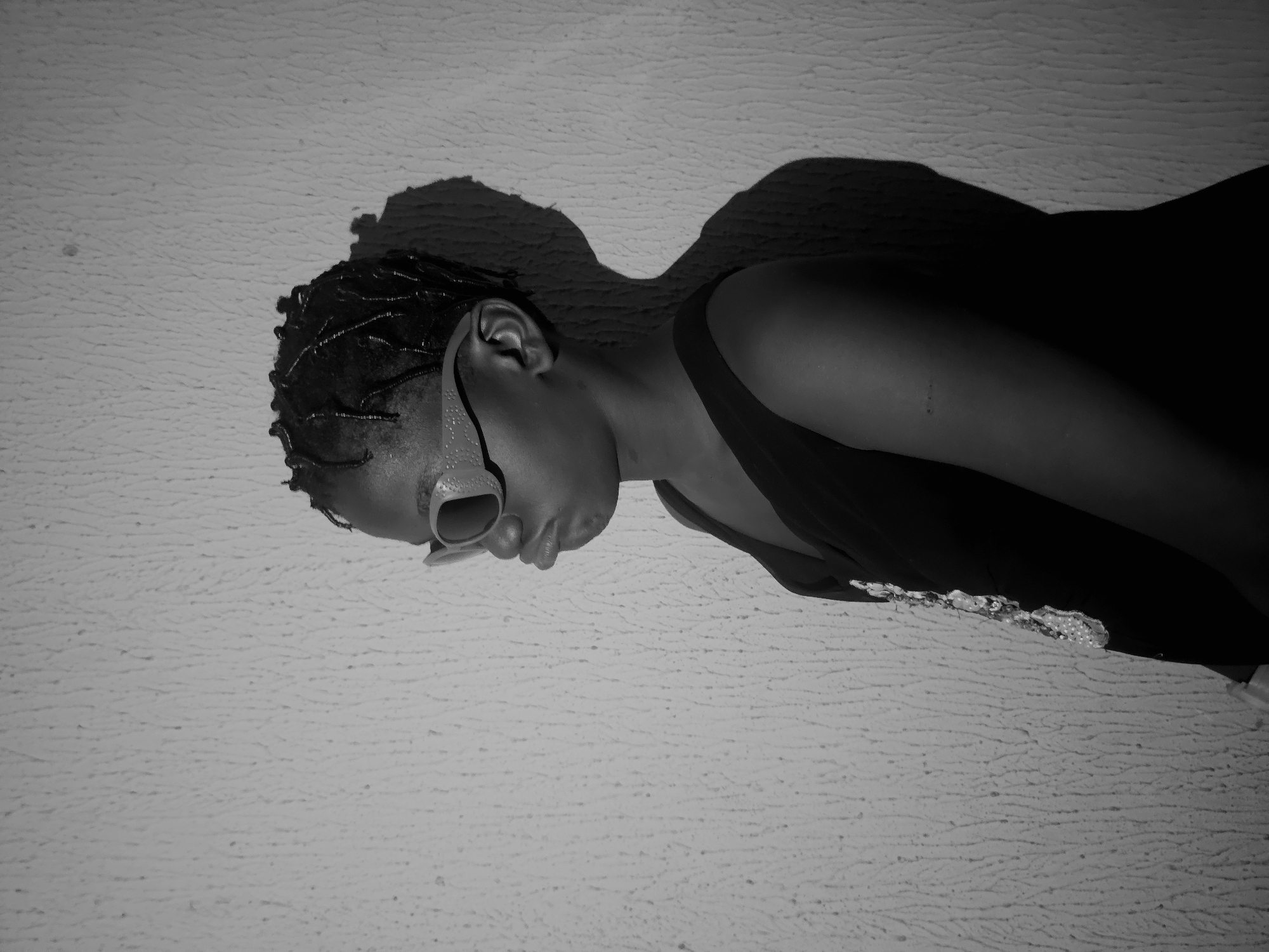 A black and white medium shot depicts a person in an Asaba, Nigeria swimming pool. The person lies on their side at the edge of the pool. The person is wearing goggles and a dark bathing suit. The person's shadow is visible on the concrete surface next to the pool. The surface has a slightly rough texture.