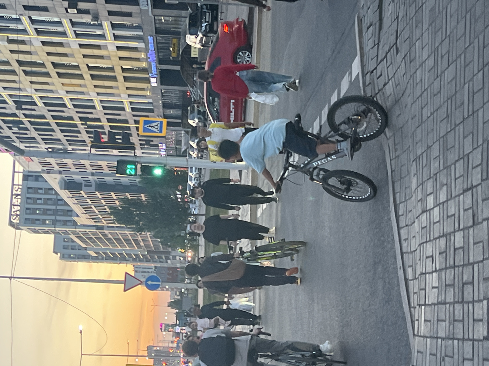 A street scene in Astana, Kazakhstan, shows people crossing a road near a modern building. A traffic light displays a green signal, and a pedestrian crossing sign is visible. Several individuals are walking across the street, some with bicycles. One person is riding a bicycle in the foreground. The background features tall, contemporary buildings and vehicles, suggesting an urban environment. The sky indicates it is late afternoon or early evening.