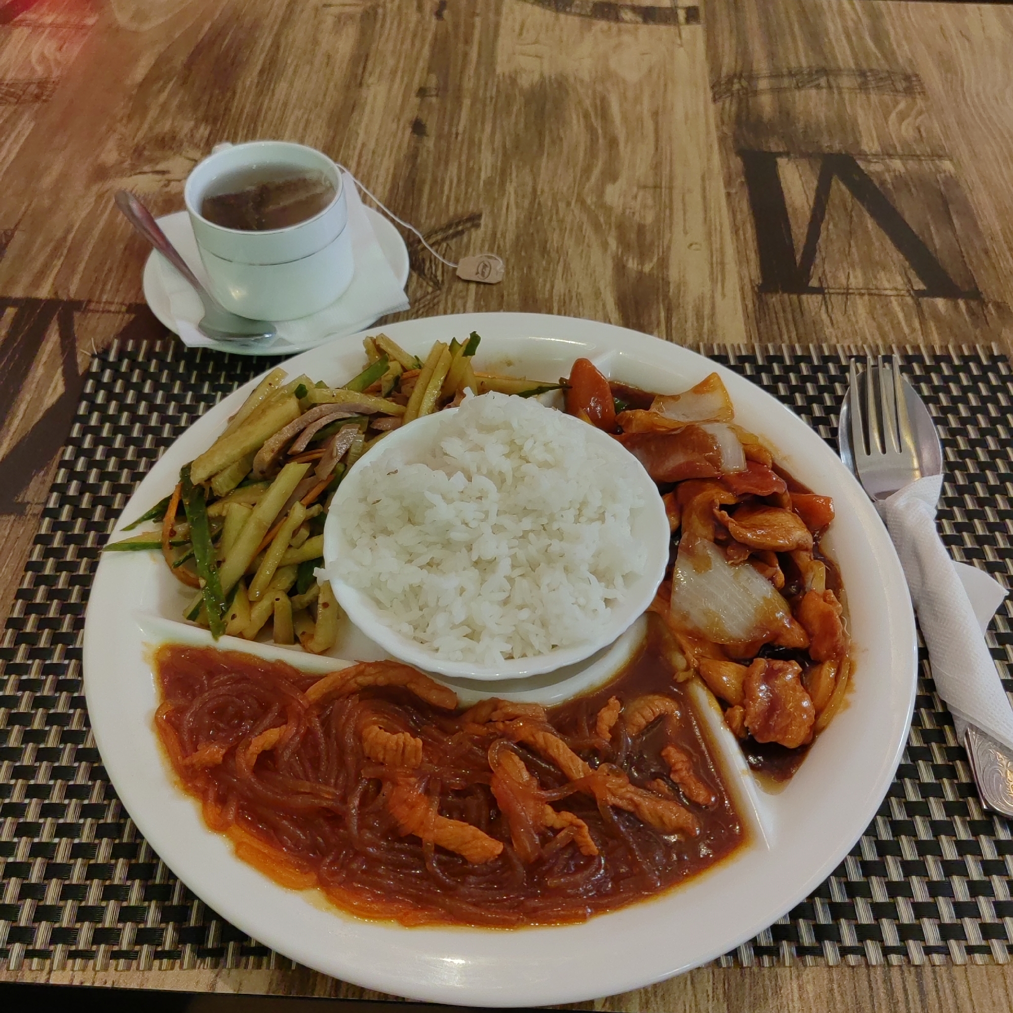 A close-up, eye-level shot captures a segmented white plate filled with a meal and a cup of tea. The plate is divided into three sections. The top left section contains a salad of thinly sliced vegetables, possibly cucumber and carrot, with slivers of meat. The top right section holds stir-fried chicken pieces in a reddish-brown sauce with chunks of white onion. The bottom section features noodles in a thick, dark sauce with pieces of meat. A small white bowl of white rice is placed in the center of the plate. To the left of the plate, a white teacup with a silver rim sits on a saucer, containing tea and a tea bag tag hanging over the side. A spoon rests on the saucer. A fork wrapped in a white napkin is positioned to the right of the plate. The meal is set on a wooden table with a textured placemat underneath the plate. The background shows a wooden surface with dark patterns, possibly a tabletop. The lighting is warm and highlights the food. No people are visible in the image. The scene is static and suggests a prepared meal ready to be eaten. There is no information in the image to associate it with Blagoveshchensk, Russia.