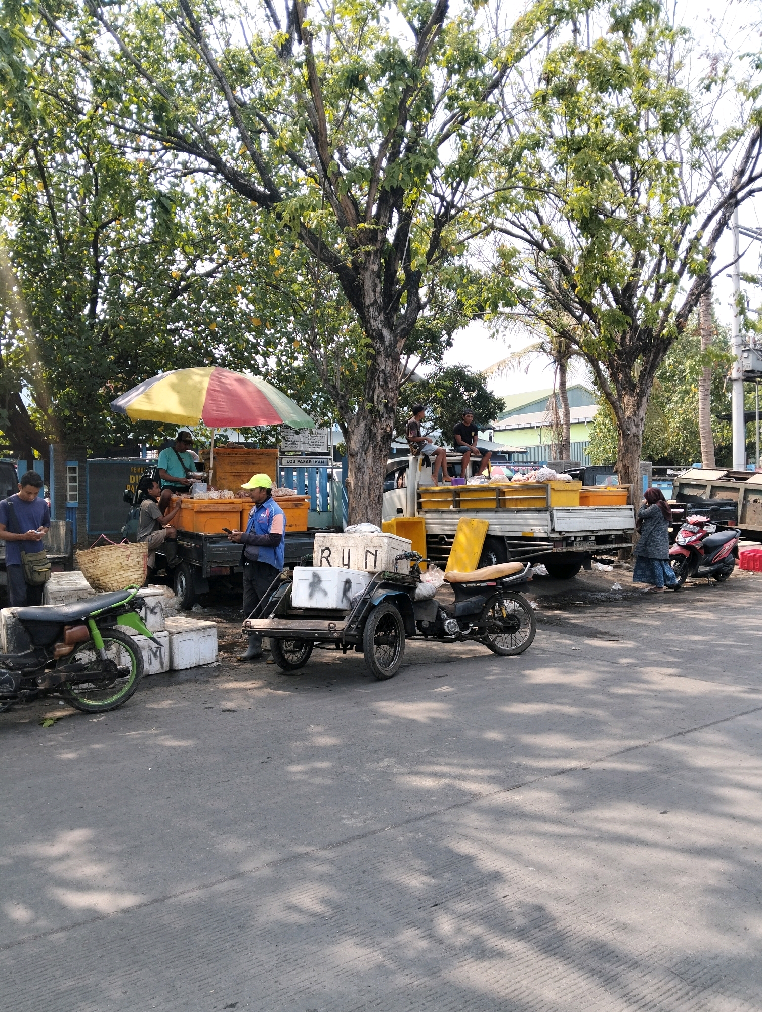 A street market scene is captured in Probolinggo, Indonesia, under bright sunlight filtering through trees. A central focus is a small truck with a yellow canopy, laden with numerous yellow crates, suggesting a goods distribution or sale. A man wearing a yellow cap and blue jacket stands beside the truck, holding a smartphone. Adjacent to the truck is a three-wheeled vehicle, possibly a motorcycle with a carriage, marked with the word "RUN" in black letters on a white cooler. A green motorcycle is parked to the left. Two men are seated on the side of the truck, and another man stands to the far left, looking at his phone. A woman in a headscarf and patterned skirt walks away from the scene towards a red motorcycle parked to the right. In the background, buildings and more trees are visible, indicating a commercial or semi-rural area. The overall impression is one of a busy, outdoor marketplace, likely on a typical day.