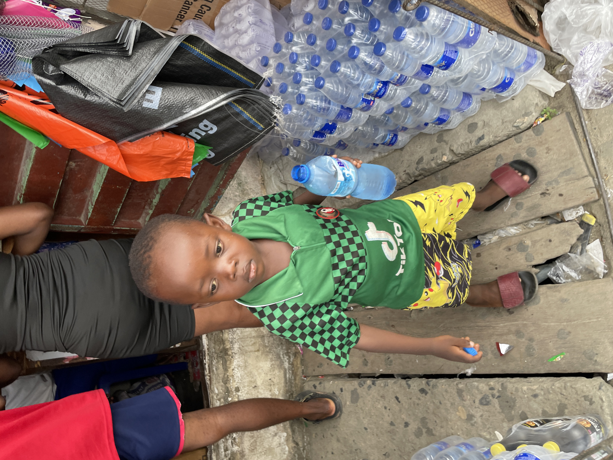 The image captures a candid moment in an outdoor setting in Ado, Nigeria, likely a market or street area. The central subject is a young child with dark skin and short, dark hair, looking directly at the camera with a neutral expression. The child wears a green long-sleeved top with a black and green checkered pattern on the sides and sleeves, and "TIKTO" (likely part of "TikTok") visible in white text on the chest. They are also wearing yellow shorts patterned with what appear to be cartoon characters, and one red slide-sandal on their right foot. The child holds a large, clear plastic water bottle with a blue cap.
To the upper right, a large stack of smaller clear plastic water bottles with blue caps and labels (partially showing "WATER") is visible. In the upper left, several large, black woven plastic bags are stacked, with the letters "ng" visible on one. An orange plastic bag or tarp is also present beneath them. A partially visible cardboard box in the background contains text that includes "Danger."
The ground is made of uneven concrete slabs or wooden planks with gaps, and scattered debris like plastic wrappers are visible. Portions of other individuals are seen to the left and bottom of the frame; one person wears a black top, and another a red top and blue shorts. The lighting is bright and even, suggesting daytime. The overall scene depicts a common daily street or market environment.