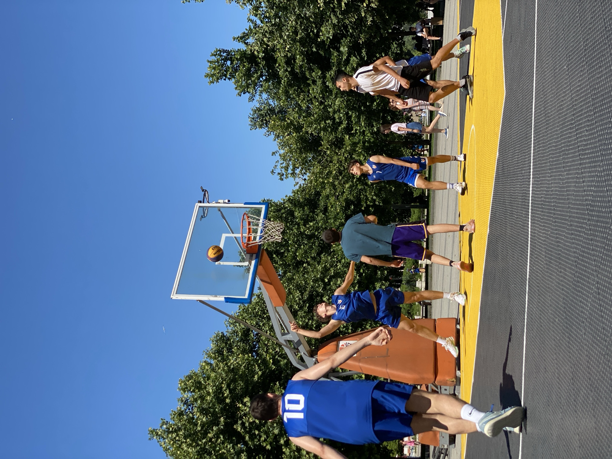 The image captures an outdoor basketball game on a sunny day, likely in Pleven, Bulgaria, given the context. The scene features a basketball court with a yellow and gray surface, a bright orange backboard support, and a clear blue sky overhead. Lush green trees form a backdrop, with a few distant figures and benches visible in the background, suggesting a park or recreational area.
Five young men are actively engaged in the game. One player, wearing a blue jersey with the number "10," is in the foreground on the left, facing away from the camera. To his right, another player in a blue jersey with "16" is mid-air, jumping to shoot the basketball. A third player in a blue jersey, with the number "7" visible, stands on the court looking up. To the right, a player in a white t-shirt and black shorts is also looking up, while another in a green t-shirt and purple shorts is positioned between the two central players. A basketball is visible in mid-air, heading towards the hoop. The players are dressed in athletic wear, including shorts and jerseys. The overall atmosphere is one of energetic sports activity in a pleasant, outdoor setting.