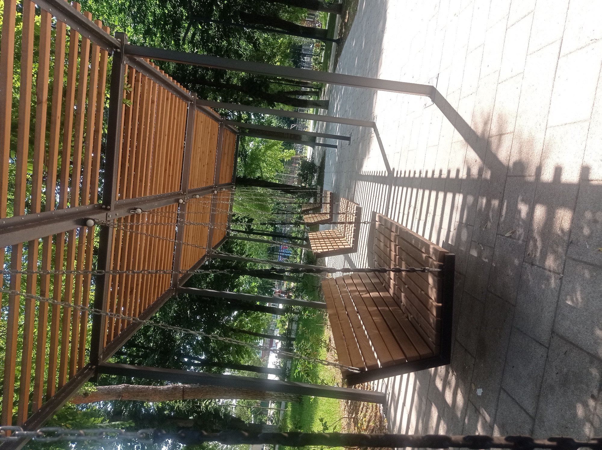A shaded area in a park features a row of wooden swings suspended from a modern, slatted wooden roof supported by metal beams and posts. The ground beneath the swings is paved with light-colored square tiles, and the area is dappled with sunlight and shadows cast by the roof and surrounding trees. In the background, lush green trees line the park, with glimpses of a grassy area and distant structures, possibly a playground. The image appears to be taken on a bright, sunny day in an outdoor recreational space.