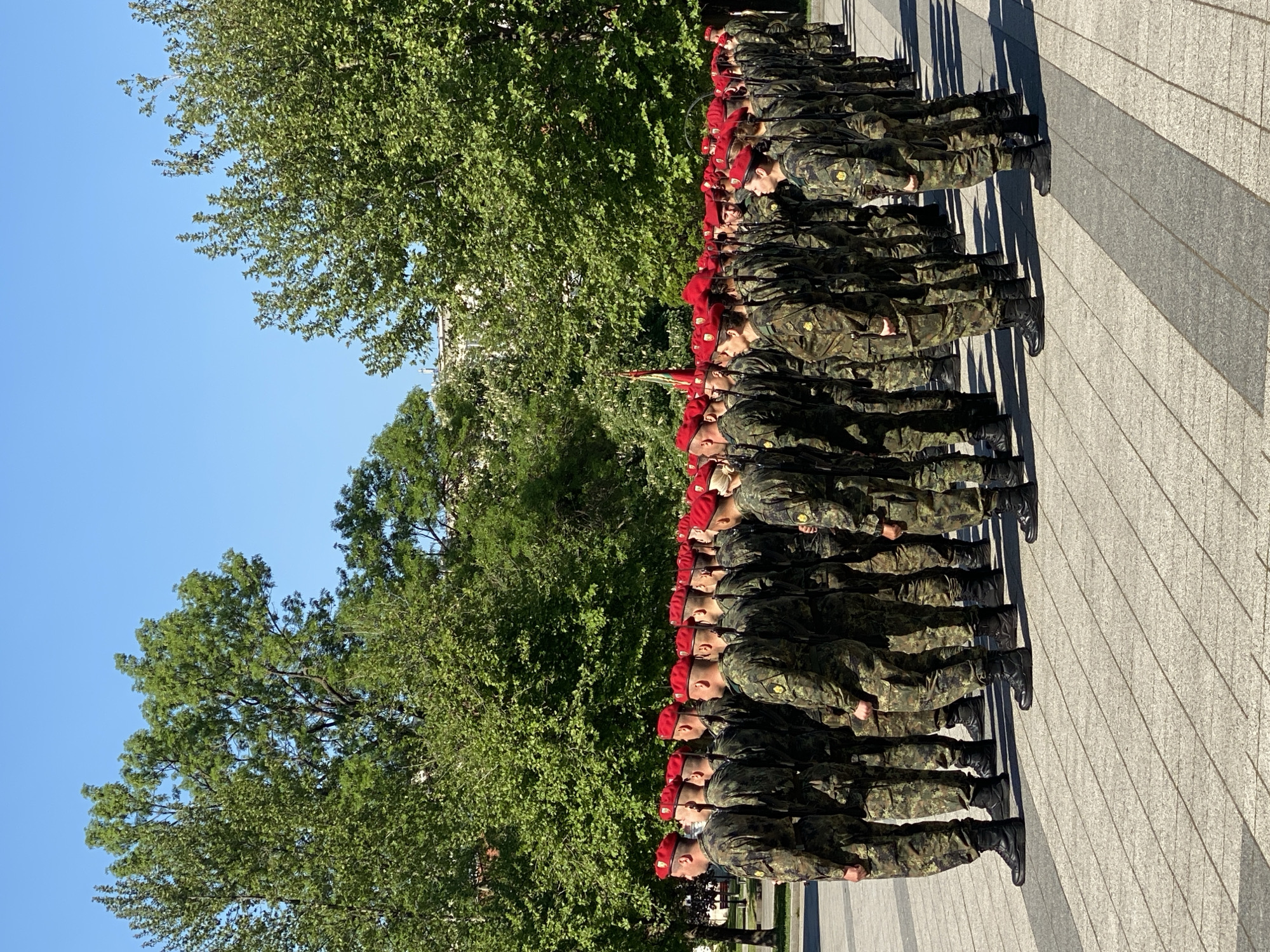 A line of soldiers in camouflage uniforms and red berets stand at attention on a stone-paved surface next to large trees. They are holding rifles. This scene is possibly located in Pleven, Bulgaria.