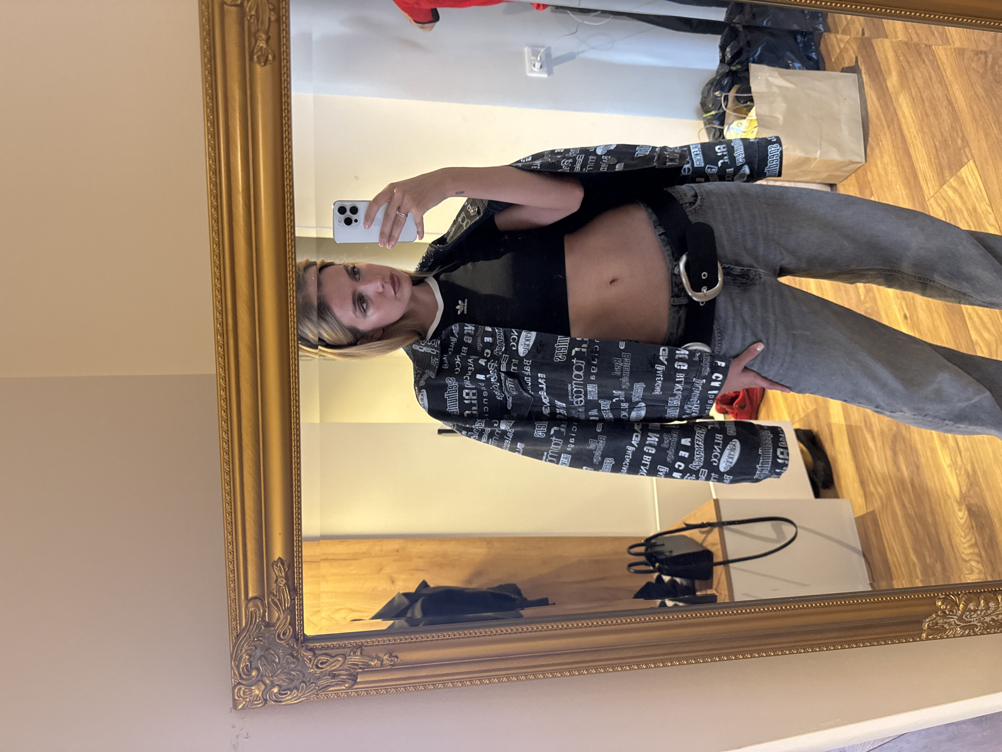 A young woman is taking a selfie in a large gold-framed mirror. She's wearing gray jeans with a wide black belt, a black crop top with white trim, and a black and white printed jacket. Her blonde hair is slightly disheveled. The background reflection shows a room with a wooden floor and a mix of items including a paper bag, a black bag, and some clothing. The walls are a neutral color. The lighting is bright, suggesting an indoor setting with natural light.