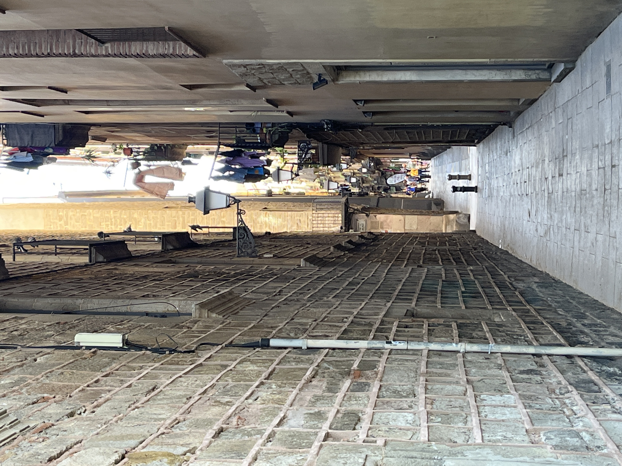 The image, when viewed in its correct orientation, depicts an upward-looking perspective from a narrow urban space, possibly a lightwell, inner courtyard, or alleyway between tall buildings.
In the foreground, a textured, tiled surface, likely a roof or lower terrace, dominates the view. This surface is composed of square-shaped bricks or tiles with prominent grout lines, appearing somewhat weathered. Several pipes and electrical conduits run across this surface, including a white rectangular electrical box on the left and a longer, cylindrical pipe extending across the width of the foreground.
As the gaze moves upward and into the distance, the facades of multiple-story buildings rise steeply on both sides, creating a deep, canyon-like effect. These buildings feature numerous windows, ledges, and balconies. Visible elements of daily life include clotheslines laden with laundry hanging from various windows and balconies, along with a few potted plants. A distinct black lamppost or light fixture extends from a wall on the left side, midway up the frame.
The scene is brightly illuminated, indicating a clear, sunny daytime, with strong light reflecting off the upper sections of the buildings. The distant background shows further architectural details and glimpses of what appear to be more buildings or upper-level structures. The dense, vertical urban environment, characterized by tall buildings and visible signs of residential life such as hanging laundry, is typical of historic European cities like Barcelona. No discernible text is visible in the image.