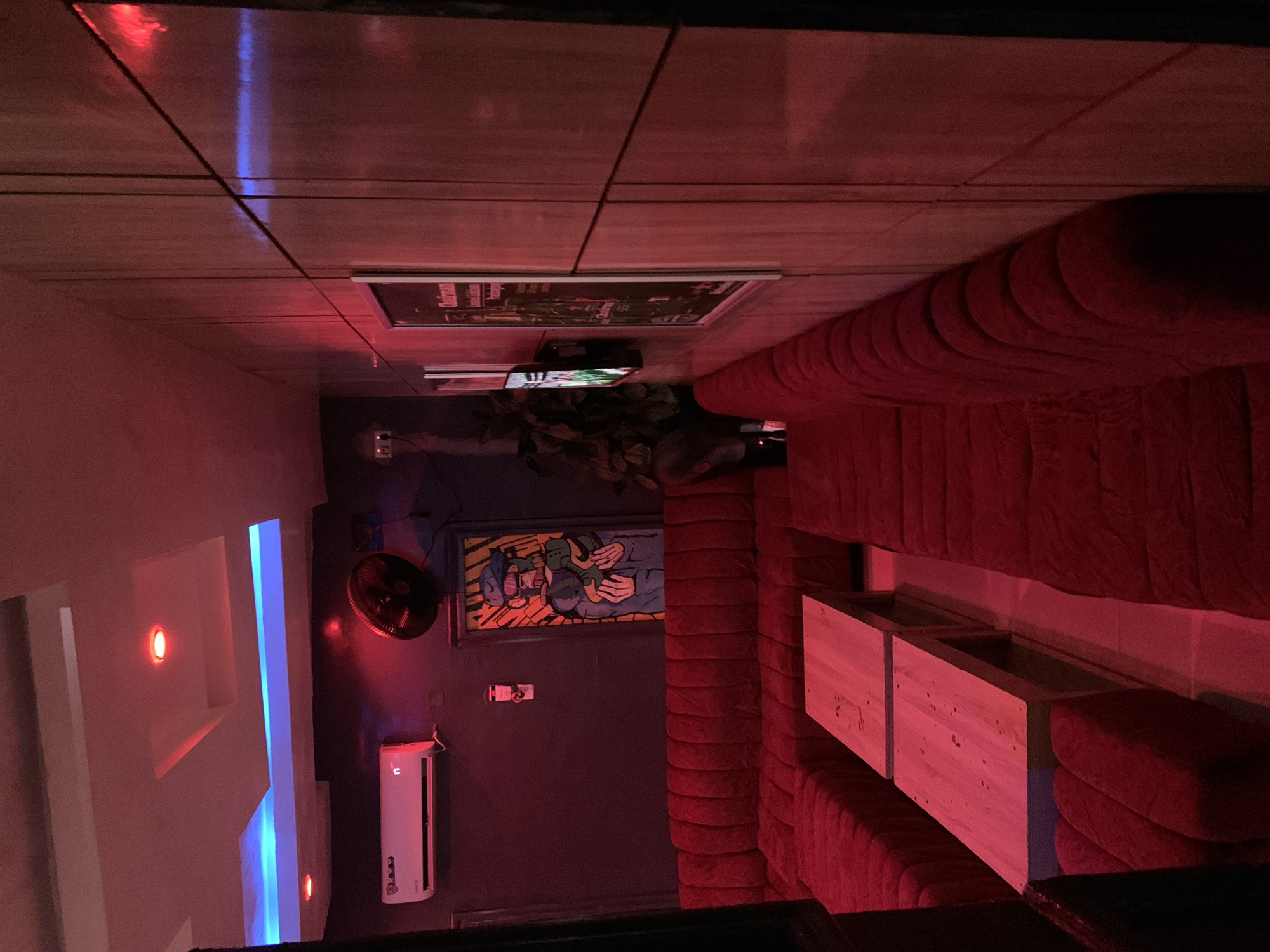 The image shows the interior of a dimly lit lounge or bar, likely in Kabba, Nigeria, decorated with red and blue ambient lighting. A long, plush red sofa with channel tufting dominates the foreground, forming an L-shape. In the center of the L-shape, a light-colored wooden table, composed of two rectangular sections, is placed on a tiled floor. The walls are paneled with vertical wooden slats on the right side of the image, and a dark-colored wall with a framed piece of art depicting a cartoonish figure in a blue jacket and hat is visible on the left. Above the sofa on the dark wall, a white air conditioning unit is mounted. A black oscillating fan is positioned above and to the right of the artwork. The ceiling features recessed lighting, with some panels illuminated in red and others with blue LED strips. On the wooden paneled wall, two framed posters are hung, one of which appears to advertise "Chairman" with some text in English. A potted plant is visible behind the seating area. The overall impression is a modern, possibly upscale entertainment venue.