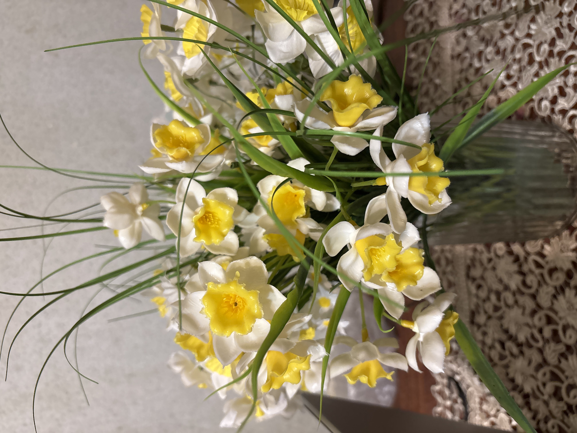 The media file displays a bouquet of artificial flowers, closely resembling daffodils or narcissus, arranged in a clear glass vase. The flowers feature white petals and prominent yellow, trumpet-shaped centers. Numerous long, thin green artificial leaves are interspersed throughout the bouquet, some curving gracefully.
The vase is made of transparent glass, showing vertical ridges on its upper portion. The floral arrangement rests on a surface covered by a light brown or beige textile with an intricate, open-work lace pattern. This patterned covering is visible on the right side of the image, extending into the background. The background behind the flowers on the left appears to be a plain, light-colored surface, possibly a wall, slightly out of focus.
No specific activity or event is taking place; the image is a static shot of a decorative floral display. The setting appears to be indoors, likely in a domestic environment. There are no discernible cues for time of day or specific weather conditions. No text is visible within the image. The image is oriented sideways, with the flowers arranged horizontally across the frame.