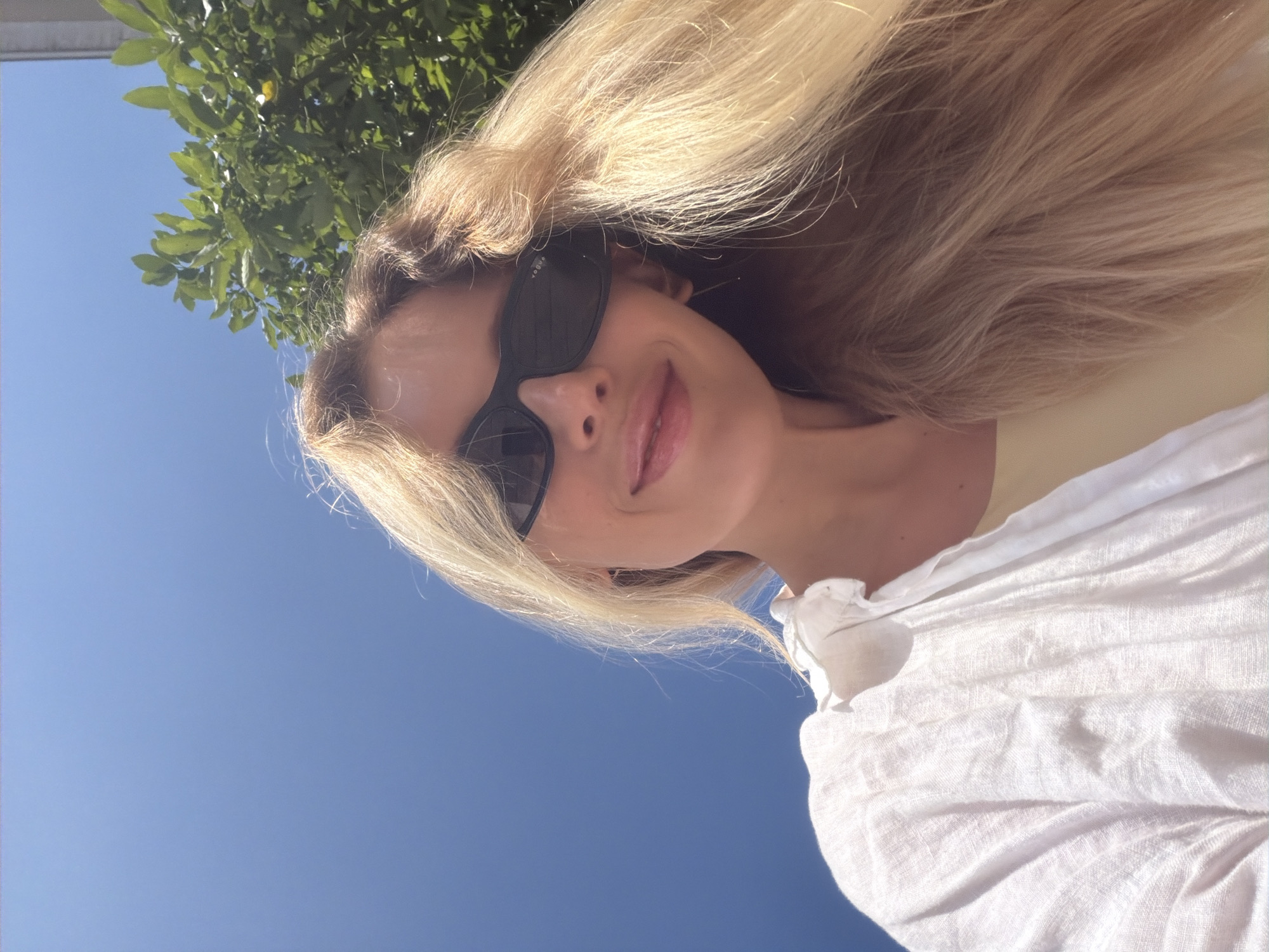 A woman is captured in a close-up, low-angle selfie outdoors. She has blonde hair, is wearing black sunglasses, and a white linen shirt. The background consists of a clear blue sky, a green tree with leaves, and a glimpse of a building. The lighting suggests it is daytime. The location is a street in the city of Budva, Montenegro.