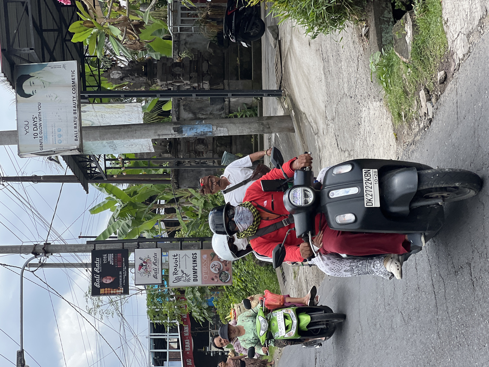 A street scene in the Ubud District of Indonesia shows three people on two motor scooters. The lead scooter is green and ridden by a woman wearing a patterned headscarf and a light green blouse. Behind her is a second person, also on the green scooter, wearing a patterned skirt. The second scooter is black and is ridden by a man in a red jacket, sunglasses, a patterned scarf over his face, and a black helmet. He is accompanied by a passenger wearing a white helmet, a white mask, and a patterned white and black skirt.
The scooters are moving along a paved road. Along the side of the road, there are several signs advertising "Bali Ratu Beauty Cosmetics," "Rabbit Gastronomy," and "Dumplings." There are also buildings, electrical wires, and lush green vegetation, including banana trees. The sky is overcast. The license plate on the black scooter reads "DK 2722 KBN." The scene appears to be daytime.