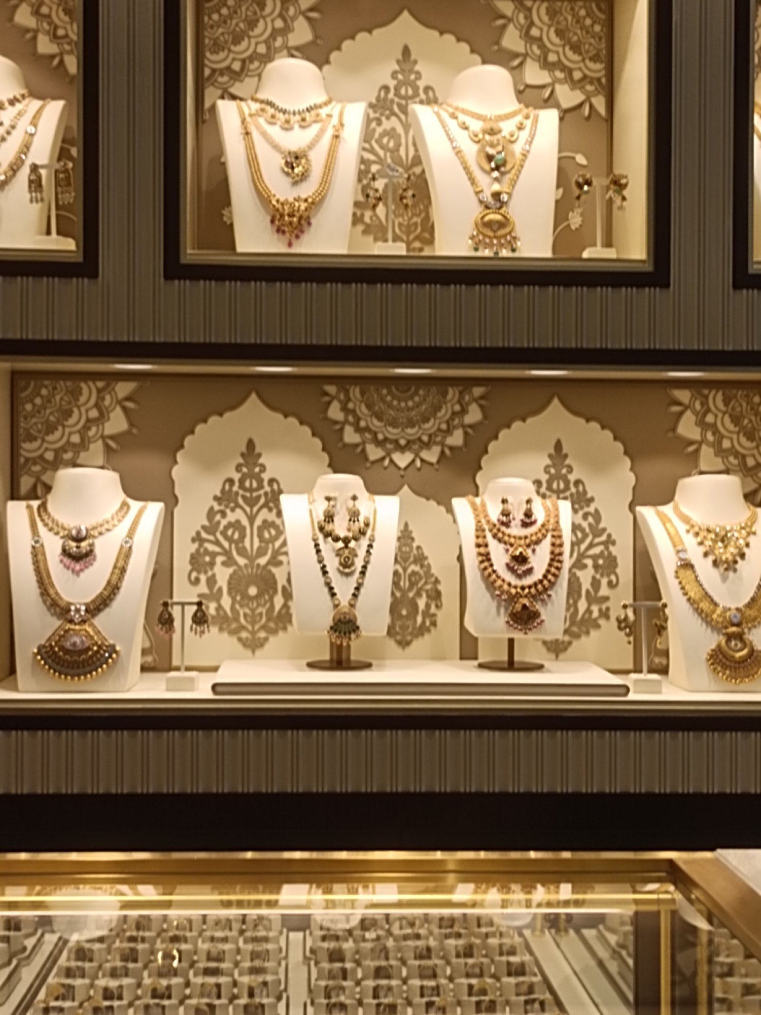 The image displays a retail display of gold jewelry in Dubai, United Arab Emirates. The display features several mannequins adorned with elaborate necklaces and earrings, primarily in gold with some gemstone embellishments. The jewelry is arranged on white busts and stands within a well-lit display case. The background of the display features decorative motifs, suggesting a high-end jewelry store. Below the main display, a glass-covered counter reveals rows of smaller jewelry items, likely rings or earrings, in individual boxes. The overall scene is one of opulent presentation common in luxury retail environments within Dubai.