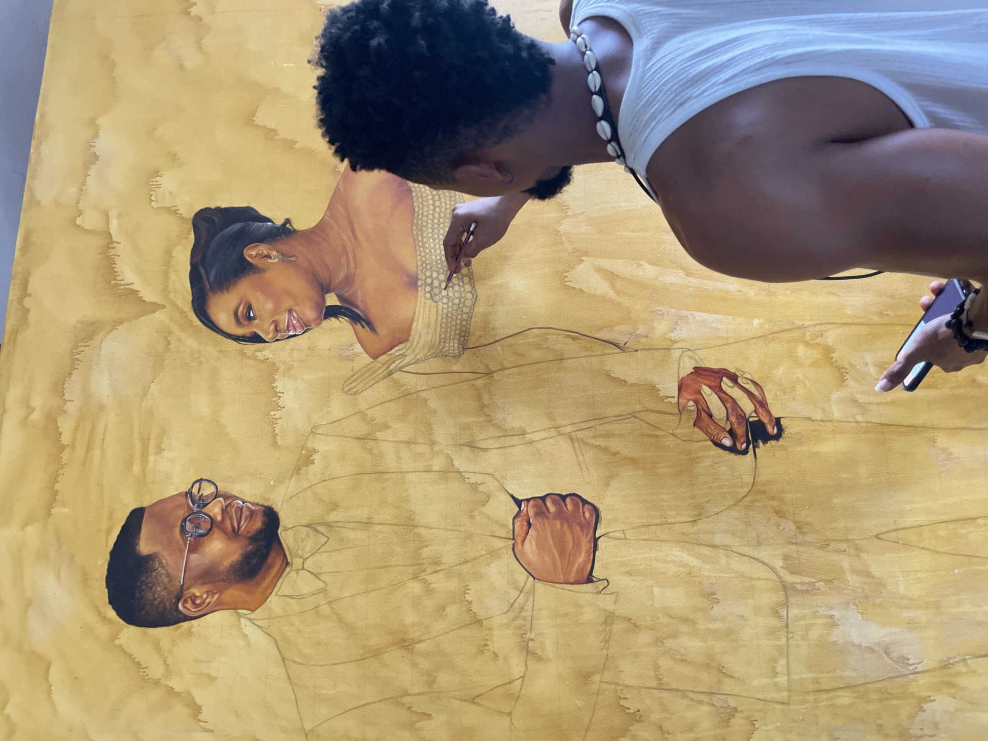 In a well-lit studio, likely located in Ado, Nigeria, an artist is captured in the process of creating a large-scale portrait on a textured, yellowish canvas. The artwork depicts a man and a woman, rendered in a realistic style with warm, earthy tones. The man, on the left, wears glasses and a suit with a bow tie, his fist clenched. The woman, to the right, is portrayed with her hair pulled back, wearing an off-the-shoulder dress. The artist, a man with dark, curly hair and a beard, stands in the foreground, facing away from the camera and towards the painting. He is dressed in a white tank top and a necklace made of white shells. He holds a paintbrush in his right hand, actively working on the woman's dress, and a smartphone in his left. The background of the studio is a muted, textured wall. The overall impression is one of focused artistic creation, with the artist deeply engrossed in his work.