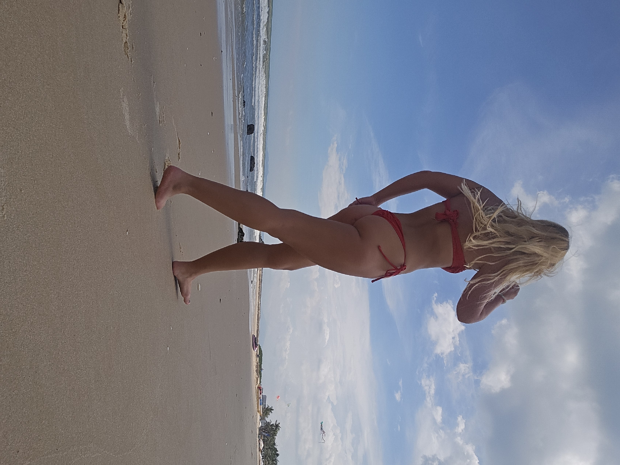 The image shows a woman in a red bikini on a sandy beach. She is standing with her back to the camera, facing the ocean. Her left leg is extended forward, and her right hand is on her hip. Her blonde hair is blowing in the wind. The ocean is calm with gentle waves, and the sky is blue with scattered white clouds. In the background, there are some rocks in the water and a distant treeline with palm trees. The beach appears to be sparsely populated, with a few umbrellas and a kite visible further down the shore. The time of day appears to be daytime, with bright sunlight casting shadows. The scene evokes a sense of relaxation and enjoyment of a beach day.