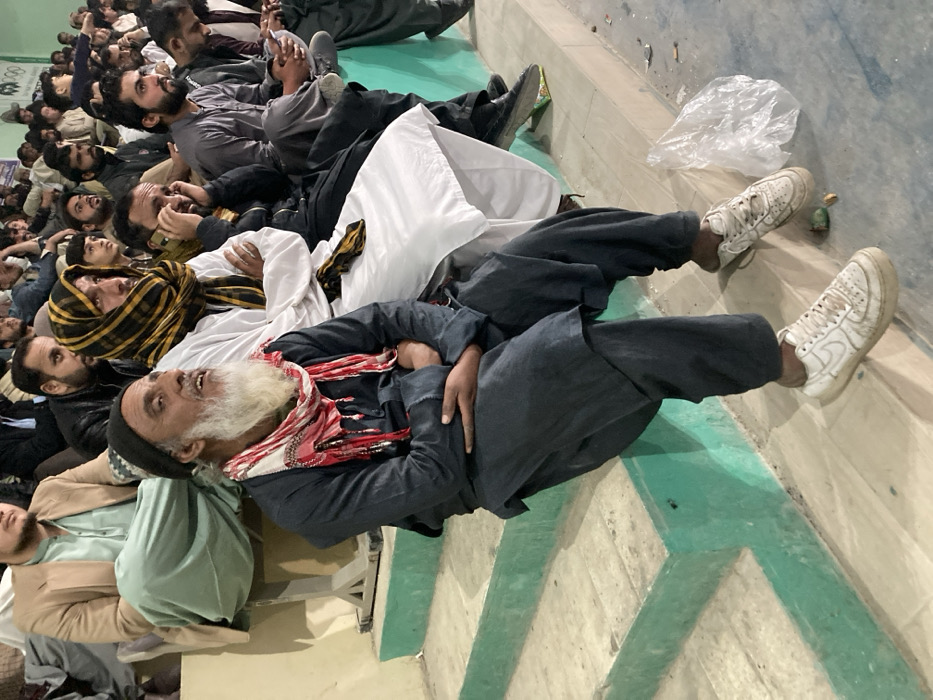 The scene depicts approximately two dozen men seated on tiered concrete spectator steps within an indoor or semi-enclosed venue in Quetta City Tehsil, Pakistan. The concrete steps feature light green painted risers and edges, with the treads being an off-white or light grey color.
In the foreground, an elderly man with a long white beard and mustache is prominently seated on a lower step. He wears a dark grey shalwar kameez, a black beanie, and a red and white patterned scarf adorned with metallic embellishments around his neck. His hands are clasped over his lower abdomen, and he looks upwards and to his right with a focused expression. He wears dirty white Nike sneakers, one of which has an unlaced tongue. To his immediate right, a man in a white shalwar kameez and a dark yellow and black patterned head covering is seated with his arms crossed, also observing an unseen activity.
Behind these two, multiple rows of male spectators are visible. Their attire includes various colored shalwar kameez, as well as blazers and jackets. One man in the mid-ground is actively engaged in a phone call, holding a dark-colored mobile phone to his right ear. Most individuals are generally looking in the same direction, suggesting observation of an event. A white wall is visible in the upper background, with a faint, partial logo resembling Olympic rings near the top right corner. On the floor level below the steps, a transparent plastic bag and a small green object (possibly a discarded fruit or wrapper) are visible among minor debris.