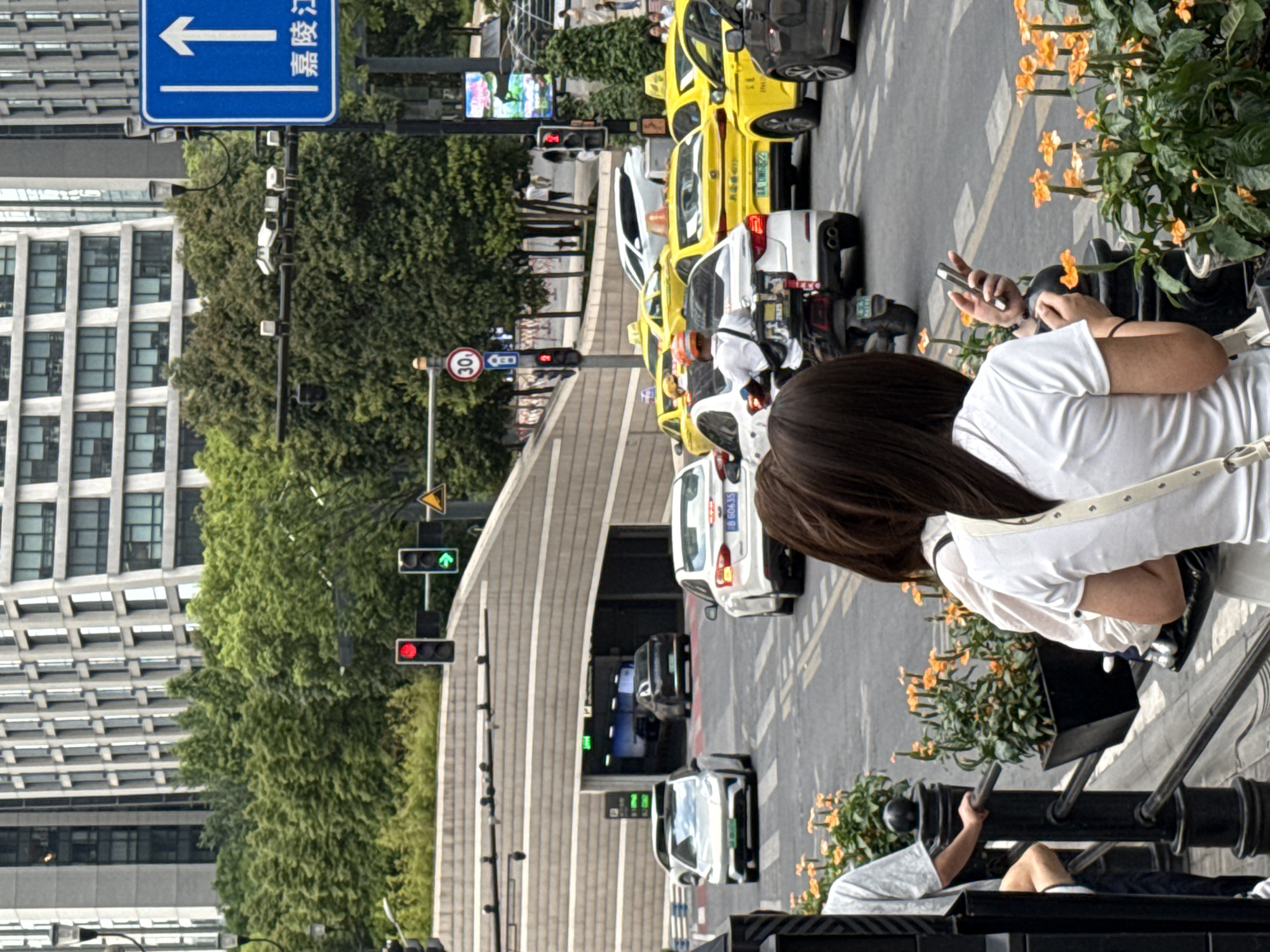 The image depicts a street scene in Yuzhong District, China, likely during daytime given the lighting. In the foreground, two women are visible from behind, one holding a mobile phone. They are standing near a black railing adorned with potted plants featuring small orange flowers.
The midground shows a busy intersection with various vehicles, including several yellow taxis with their distinctive roofs, white cars, and a motorcycle. Traffic lights are visible, with one showing a red light and another showing a green arrow. A speed limit sign indicating "30" is also present. In the background, a modern building with a patterned facade rises, alongside lush green trees. A tunnel entrance is visible on the left, with cars entering it. The street is paved, with visible lane markings.