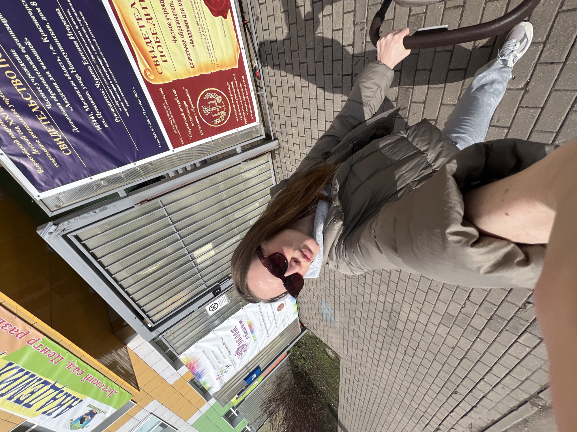 Here is a detailed description of the image:
**Scene Overview:**
The image captures a first-person perspective on a paved pathway. The primary subject is a person wearing a brown puffer jacket, grey sweatpants and sneakers, pushing a stroller. The scene is oriented toward a row of buildings and advertisements or signage.
**People:**
* The visible person is likely pushing a stroller and wearing sunglasses, a brown puffer jacket, gray sweatpants, and white sneakers.
**Subjects/Objects:**
* **Buildings:** The backdrop includes buildings with a mix of yellow and green exteriors.
* **Advertisements/Signage:** Several large advertisements are attached to the buildings, seemingly promoting a kindergarten ("Детский сад") and other educational programs. Some of these signs are written in Russian.
* **Pavement:** The walkway is constructed of gray paving stones or tiles.
* **Stroller:** A stroller frame is visible.
**Actions/Interactions:**
* The person is actively pushing the stroller along the paved walkway.
**Environment:**
* **Location:** Based on the Russian text in the advertisements, the location is likely within Russia. It is located in the city of Krasnogorsk, near Moscow.
* **Weather/Lighting:** The scene appears to be outdoors with bright sunlight.
In summary, the image shows a person strolling a stroller along a paved pathway in Krasnogorsk, Russia, with buildings and advertising signage in the background under bright sunlight.