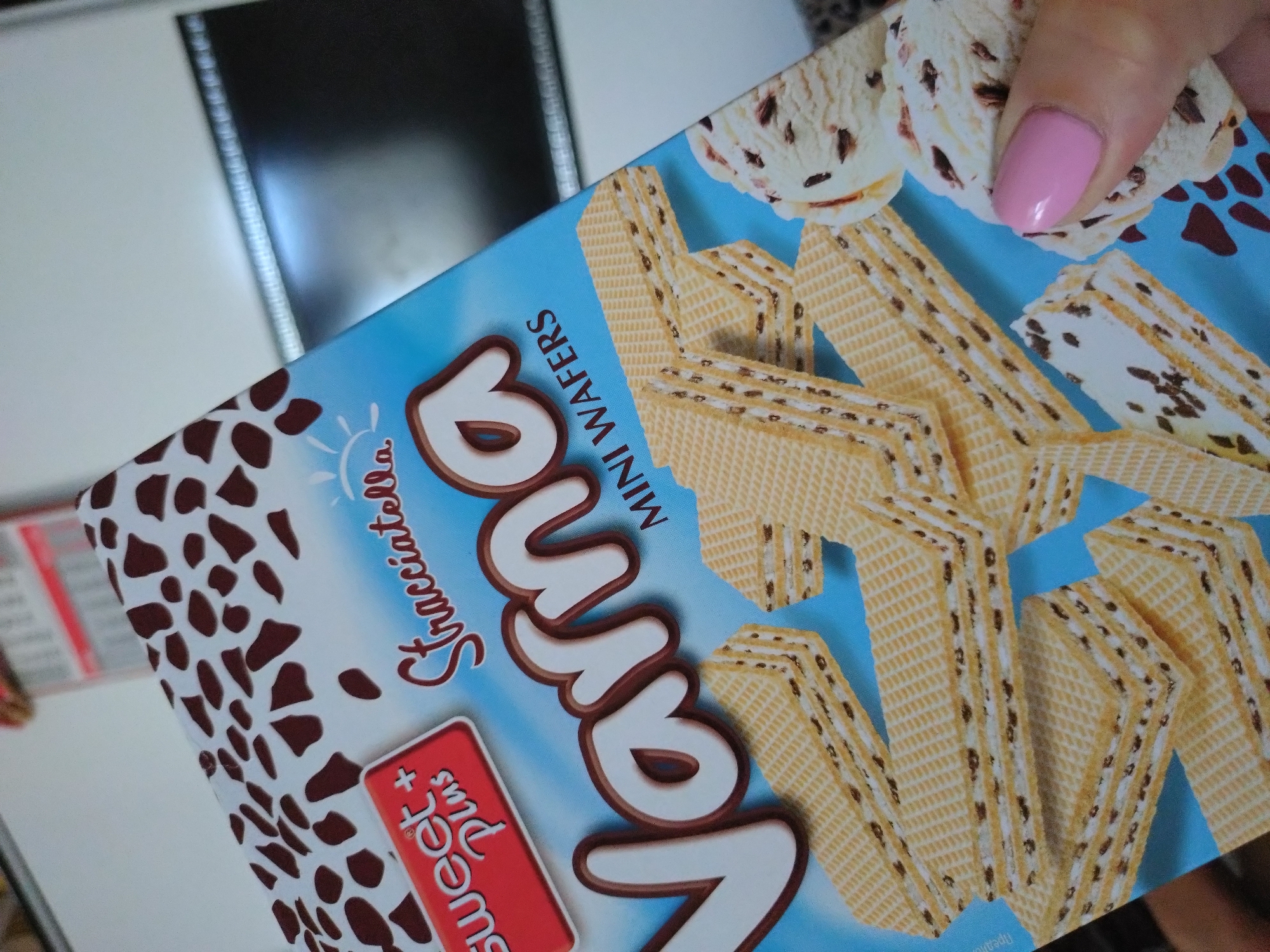 The image shows a close-up of a box of Varna brand mini wafers with a stracciatella flavor. The box is light blue with a pattern of dark brown chocolate-like splatters on the upper portion. Text on the box includes "sweet + Plus" in a red rectangle, "Stracciatella" in elegant script, and "Varna" in large, white, stylized lettering. Below the "Varna" text, "MINI WAFERS" is printed in smaller, dark letters. The foreground features several stacked mini wafers, which are rectangular and have a textured surface with visible layers of cream filling and dark chocolate chips. To the right, a hand with a pink fingernail is holding a scoop of ice cream, which appears to be vanilla with dark chocolate pieces, similar in appearance to the wafers. The background is blurry but suggests an indoor environment, possibly a store or a kitchen, with a white surface and a dark rectangular object, likely a television screen or monitor. There is no discernible indication of the specific location within the city of Stara Zagora, Bulgaria.