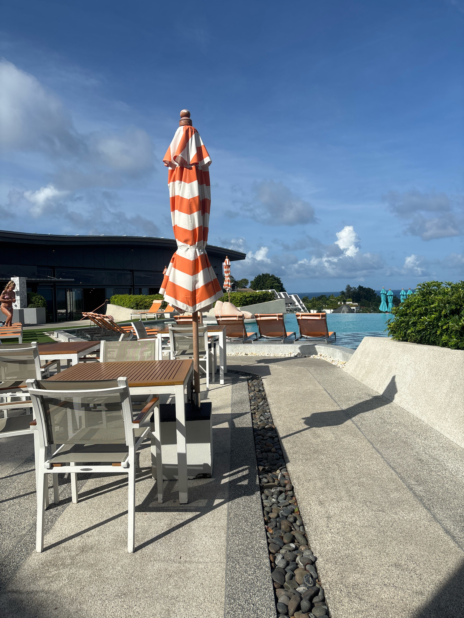 The image depicts an outdoor seating area at a resort in Karon, Thailand. In the foreground, several tables and chairs with white frames and wood-like tops are arranged on a paved surface. An orange and white striped umbrella, partially closed, stands prominently in the center of the frame. To the right, the shadow of the umbrella falls across the pavement.
In the mid-ground, lounge chairs are visible around an infinity pool that overlooks the ocean. Several turquoise umbrellas are lined up near the pool's edge. A modern building with dark windows and a dark roof forms the background. To the left of the building, a person in a bikini can be seen standing on a bench. The sky above is blue with scattered white clouds. The overall scene suggests a sunny day at a tropical resort.