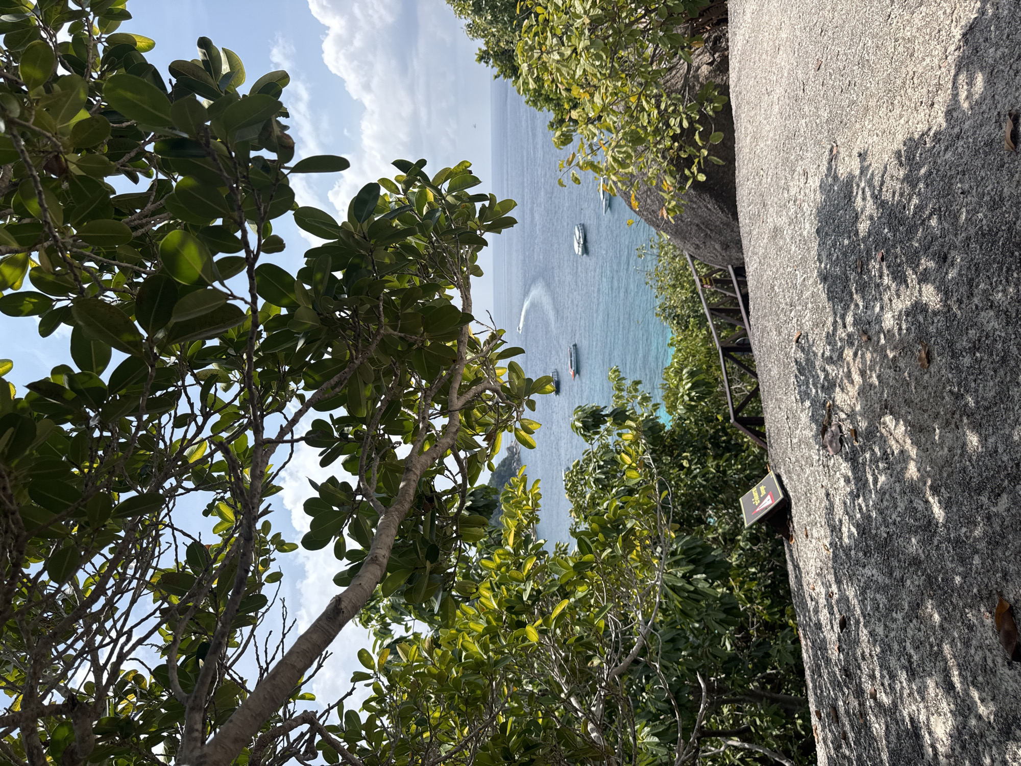 The image shows a coastal view from a high vantage point in Lam Kaen, Thailand. The foreground is dominated by lush green foliage of trees and shrubs. In the center of the frame, boats are visible on the blue water, and a white water trail indicates a vessel in motion. A large gray rock formation with a warning sign on it is on the right, with shadows cast upon its surface. The sky is blue with scattered white clouds.
