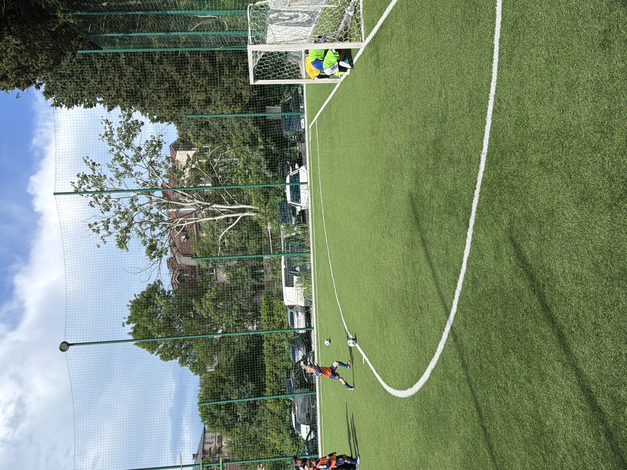 The image depicts a soccer game in progress on an artificial turf field. A player in an orange and blue uniform is diving towards two soccer balls near the edge of a white line marking part of the playing area. Other players are partially visible at the bottom edge of the frame, also in orange and blue uniforms. A goal post is visible on the far side of the field, with a person in a green, yellow, and blue uniform inside. A green net runs along the side of the field, behind which are trees, buildings, and parked cars. The sky is blue with clouds. The location is Sochi, Russia, based on visual clues of urban development and the presence of the Caucasus Mountains in the background.