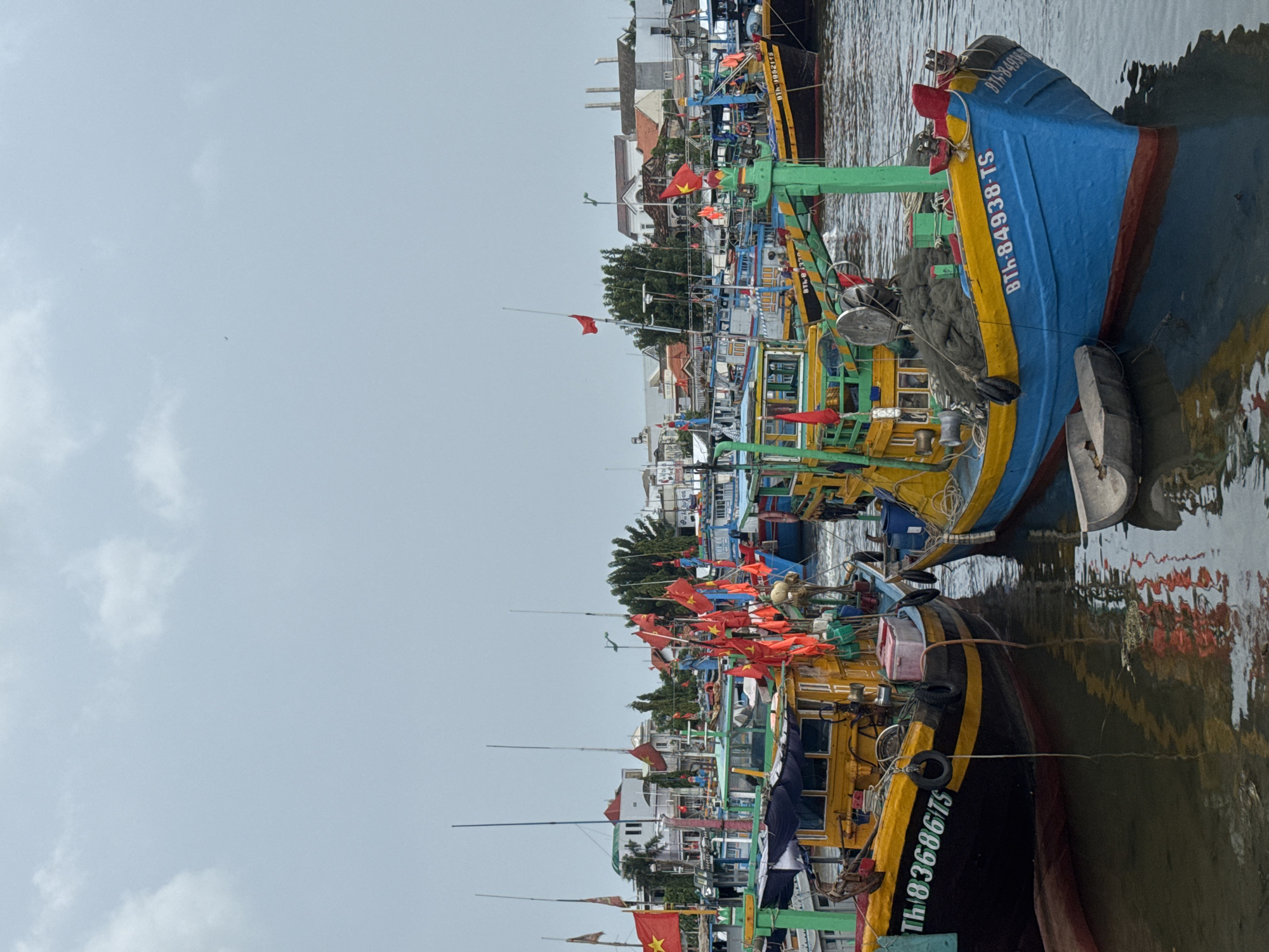 The image depicts a harbor in Phan Thiết, Vietnam, filled with numerous fishing boats. The sky is overcast with light clouds, and the water is calm, reflecting the boats and flags. The boats are predominantly colorful, with many adorned with red Vietnamese flags. Some boats are docked and appear to be of various sizes and designs, typical of fishing vessels in the region. In the background, buildings and trees are visible, suggesting a coastal town or city. The overall scene conveys a sense of activity and maritime life.