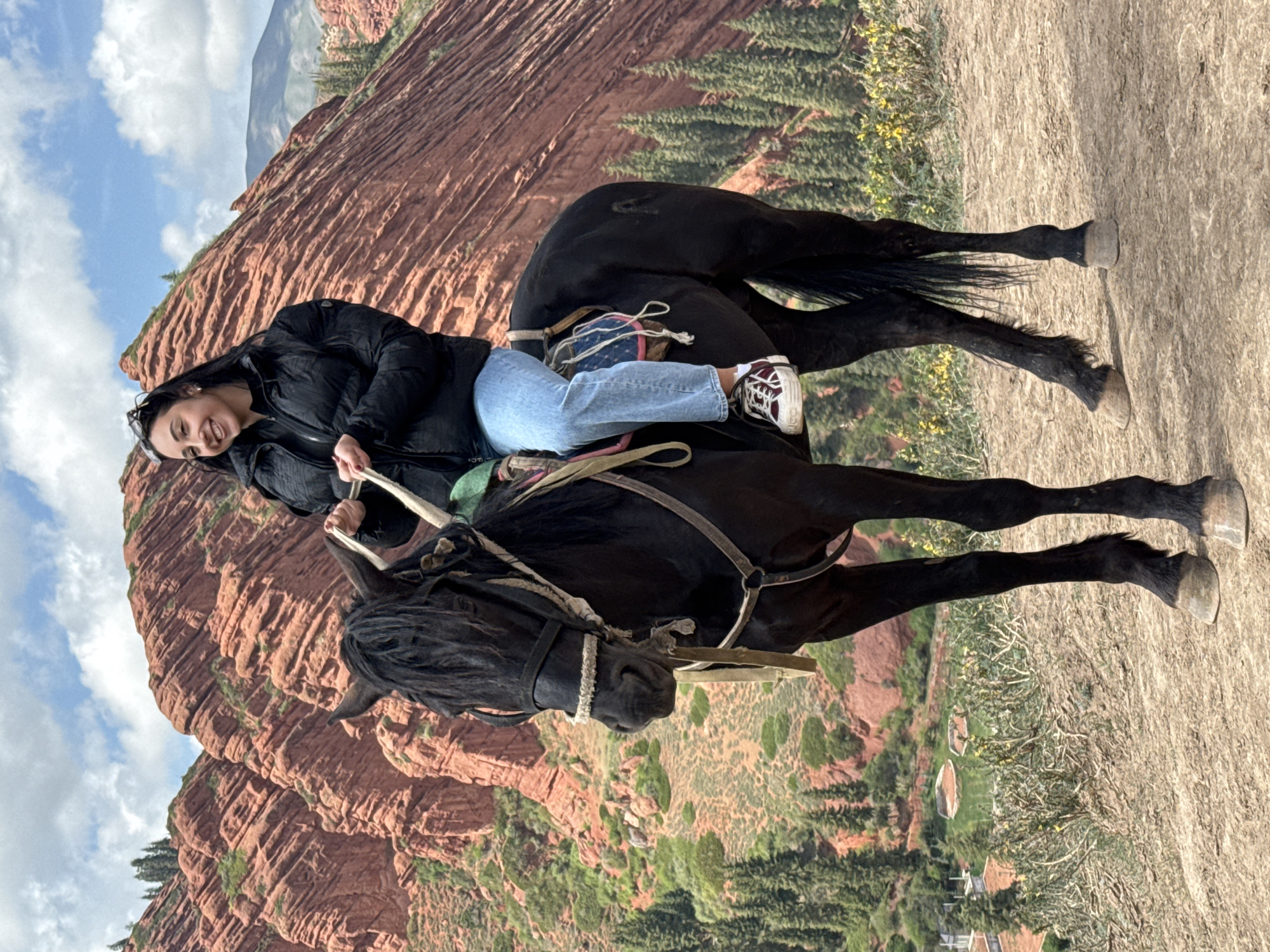 A vibrant, daytime image depicts a young woman horseback riding through the striking red rock landscape of Jeti-Ögüz, Kyrgyzstan. The woman, who appears to be in her 20s or 30s, is seated on a black horse. She is wearing a black puffer jacket, light blue jeans, and maroon and white sneakers, with sunglasses resting on her head. Her long dark hair frames a wide, happy smile as she looks towards the camera, holding the horse's reins.
The black horse is equipped with a bridle and a simple saddle, and appears to be walking on a dusty path or terrain. In the background, towering, layered red sandstone formations dominate the scene, characteristic of the "Seven Bulls" rocks in the region. Green patches of trees and vegetation are scattered across the mountainous terrain, contrasting with the rich red earth. The sky is partly cloudy with bright white clouds against a blue backdrop, indicating clear and pleasant weather conditions. The overall atmosphere conveys joy and enjoyment of the outdoor activity and scenic environment. There is no visible text in the image.