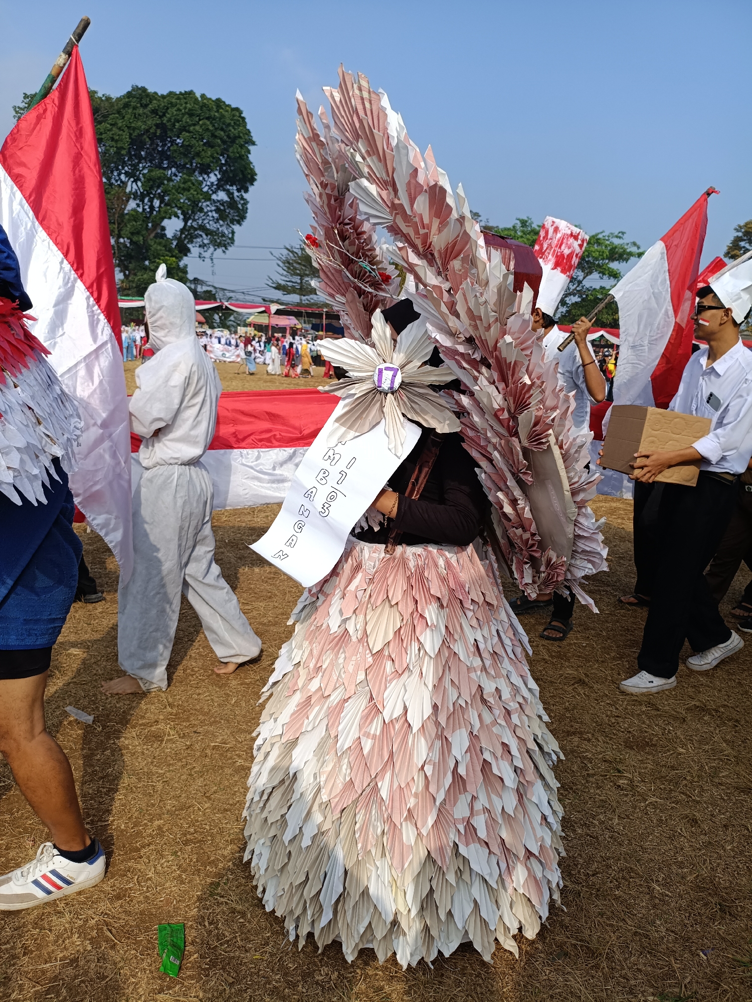 The image depicts an outdoor event, likely a parade or celebration, taking place on a dry, grassy field. The sky is clear and blue. In the foreground, a person in a costume made of layered, folded paper in shades of pink and white stands prominently. The costume has a large, elaborate headdress or shoulder piece constructed from similar folded paper, resembling feathers or wings. A white banner with text in black is attached to the costume.
To the left of this central figure, an Indonesian flag (red and white) is visible, attached to a wooden pole. Further to the left, a person wearing a blue garment and white sneakers is partially visible. Behind the central figure, a person in a white hazmat suit is walking, carrying a large red and white flag.
In the background, there are other participants, some holding flags, and tents or structures that suggest a public gathering. The overall atmosphere appears festive and communal. The presence of Indonesian flags strongly suggests the event is related to Indonesian national pride or a national holiday. The context provided indicates the location is Cibodas, Indonesia.