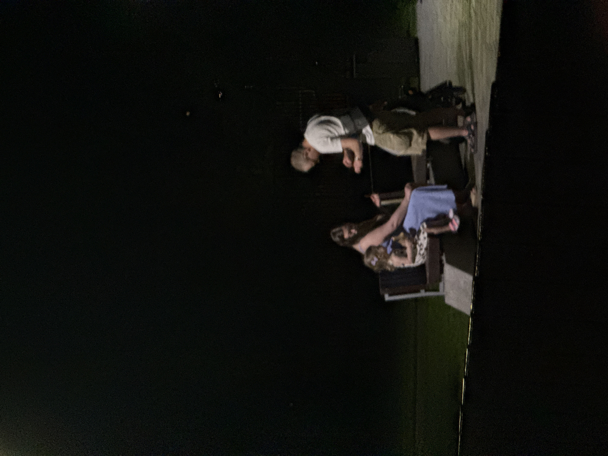 The image depicts an outdoor scene at night, where three individuals are visible. A woman, identified as an adult, is seated on a bench, holding a child, also an adult, on her lap. The child is wearing a patterned dress and has a hair accessory. To the right of the bench, a man, also an adult, stands, facing the woman and child. He is wearing a white t-shirt and khaki shorts. He appears to be engaged in a conversation or interaction with the woman. The background is dark, with some faint illumination suggesting an outdoor setting. There is a bench and what appears to be a bicycle or scooter behind the man. The location is specified as the city of Dnipro, Ukraine.