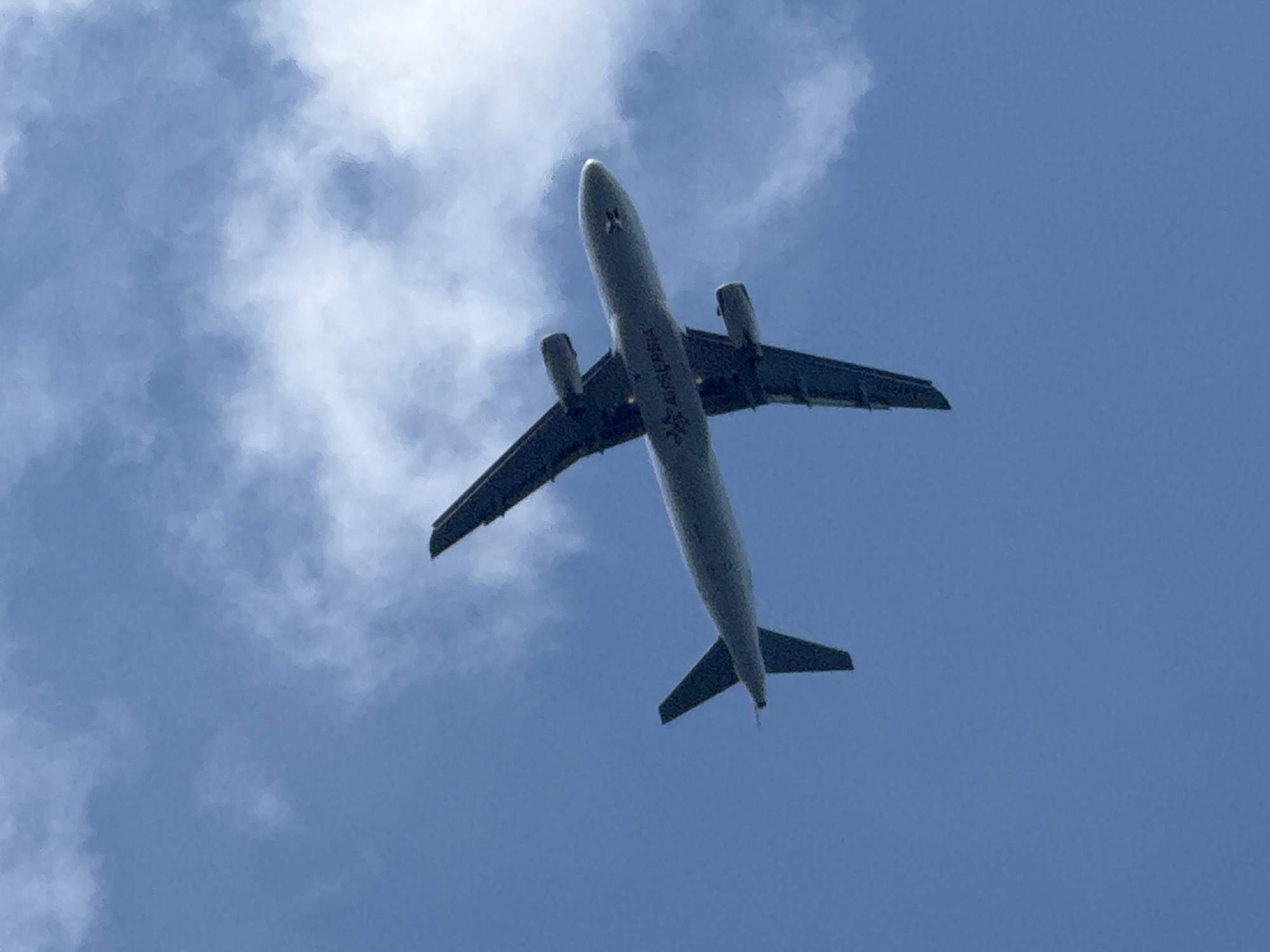 An airplane is viewed from below, flying against a clear blue sky with scattered white clouds. The underside of the airplane is visible, with its wings extended to the sides and its tail fin pointing upwards. The airline's logo, which appears to be "AIR SERBIA," is visible on the fuselage. The airplane is captured mid-flight, suggesting it is either taking off or landing. No specific location in Budva, Montenegro is identifiable from the image.