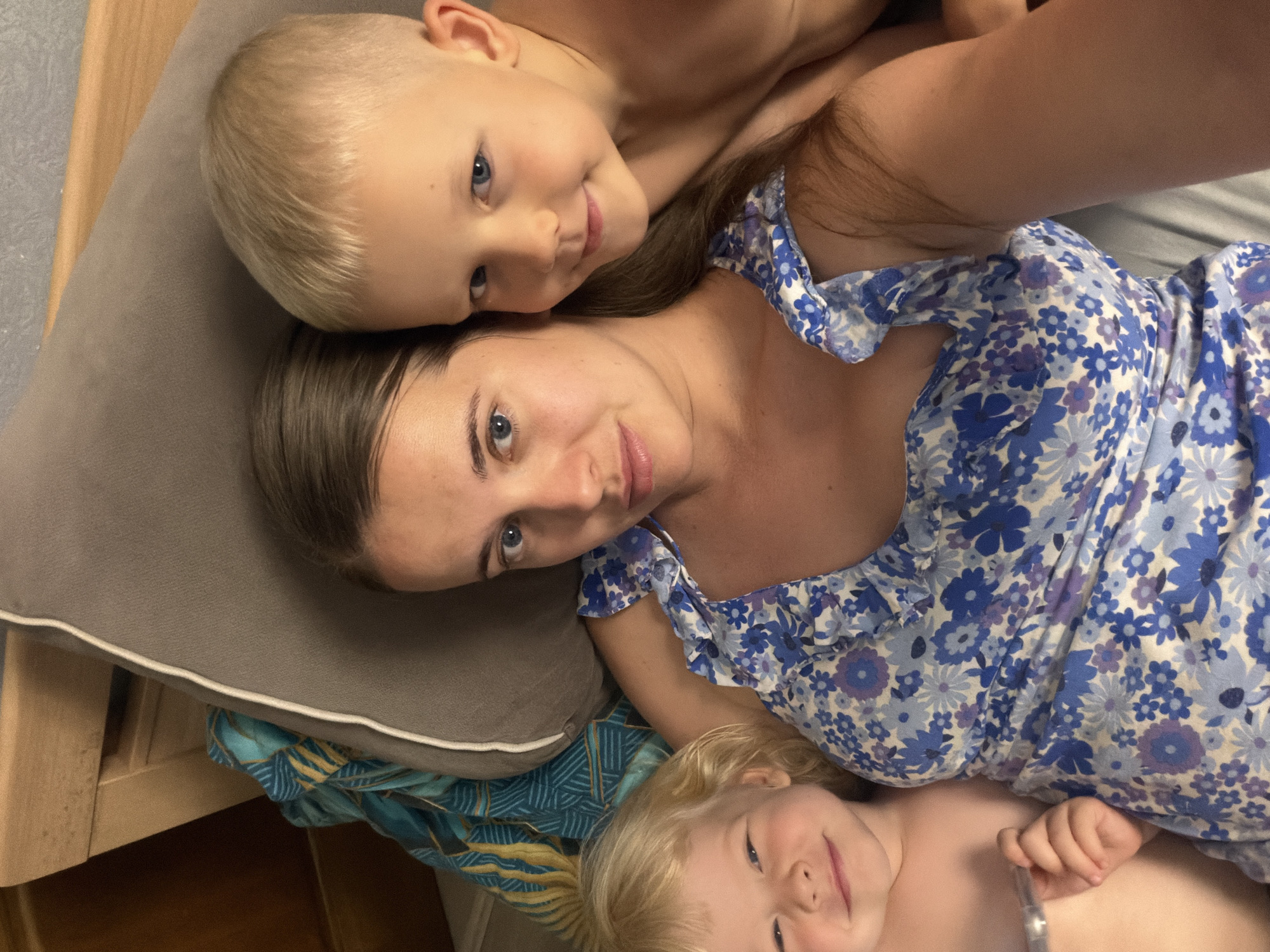 A top-down, slightly angled close-up selfie shows a woman with two children, likely taken indoors in Sochi, Russia. The woman, with fair skin, brown hair, and blue eyes, lies on her back, looking directly at the camera with a soft expression. She is wearing a blue and white floral-patterned dress.
Above her head, a young boy with blond hair and blue eyes rests his head on her, also looking at the camera with a gentle smile. He appears to be shirtless. Below the woman, a younger child with blond hair and blue eyes is visible, smiling and looking up towards the camera. This child is also shirtless, and a small hand can be seen near the woman's dress.
The subjects are positioned on what appears to be a bed or sofa. A gray pillow is visible under the woman's head, and patterned blue and yellow fabric can be seen beneath her and the younger child. A light wooden headboard or furniture piece is partially visible on the left side of the frame. The overall atmosphere appears calm and affectionate. No discernible text is visible in the image.