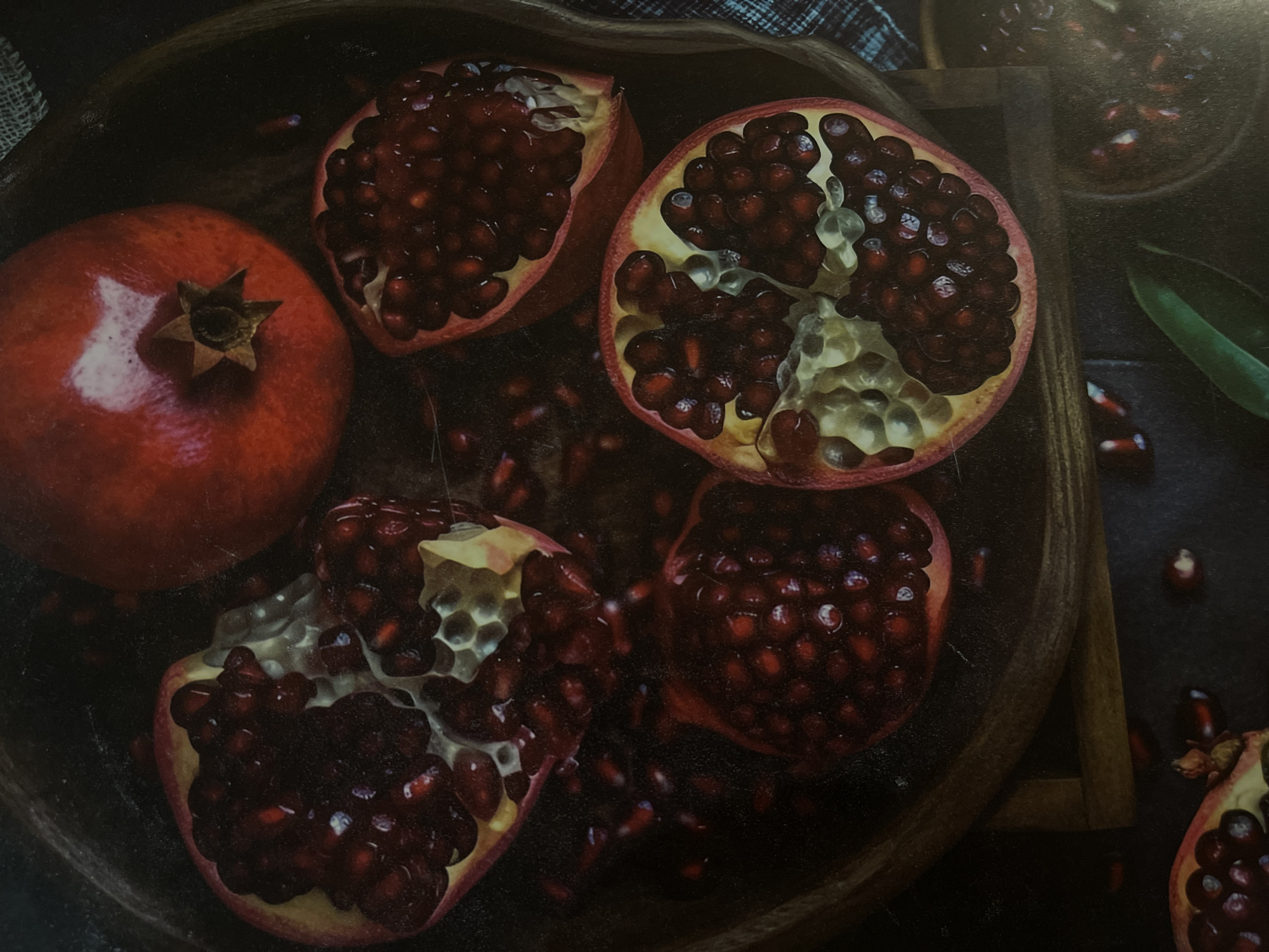 The image displays a close-up, top-down view of several pomegranates arranged in a wooden bowl. One whole pomegranate rests at the top, while several others are cut open, revealing their ruby-red arils. Some arils have spilled out around the fruits. The lighting is dim and moody, highlighting the textures and colors of the pomegranates. A blue and white checkered cloth is visible in the background. The scene is static and appears to be a still life arrangement. There is no discernible location within the city of Minsk, Belarus, or any indication of it being in that city.